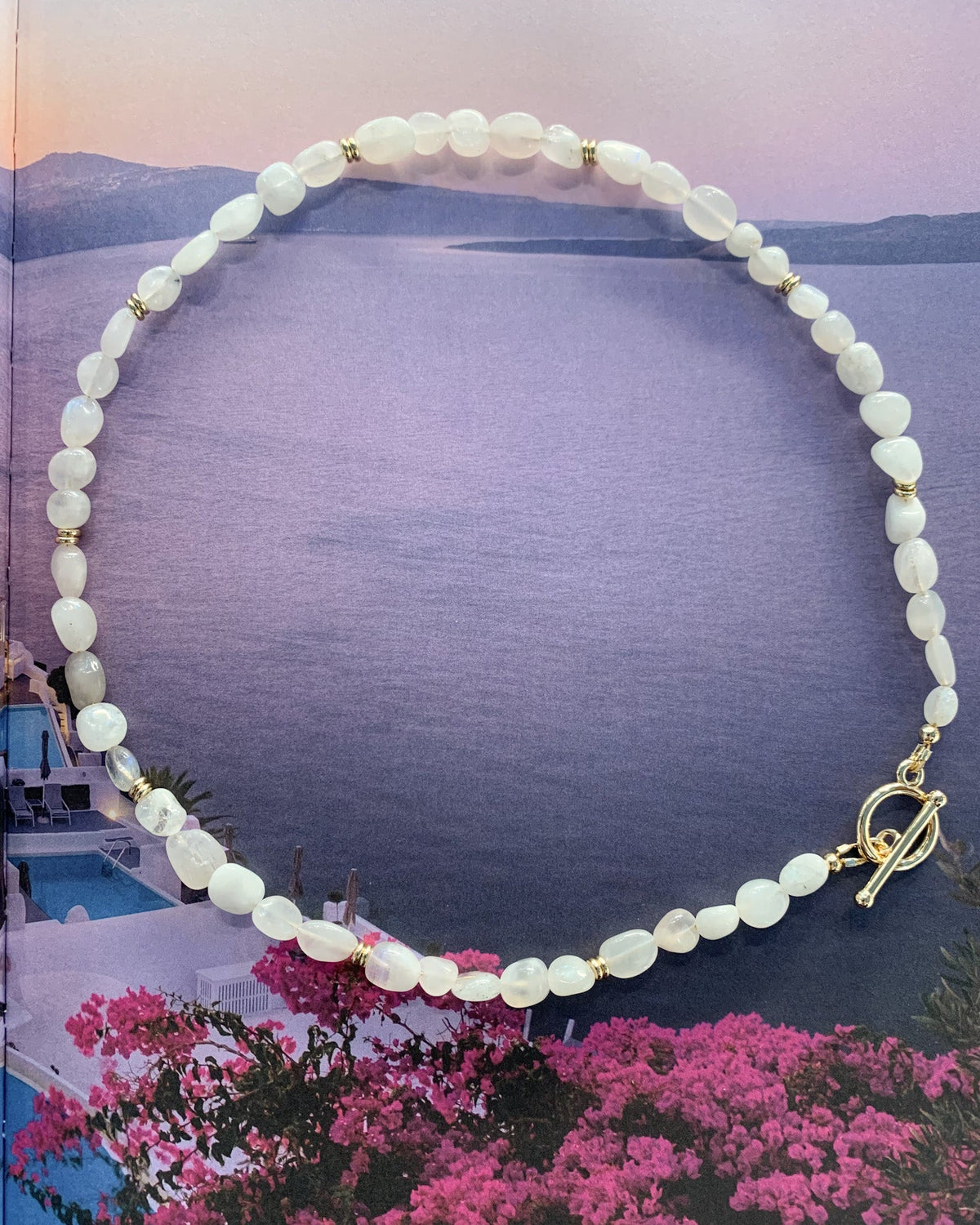 A beaded necklace made of white oval moonstone beads with small gold spacer beads and a gold toggle clasp, displayed over a scenic background featuring purple water, mountains, and pink flowers.