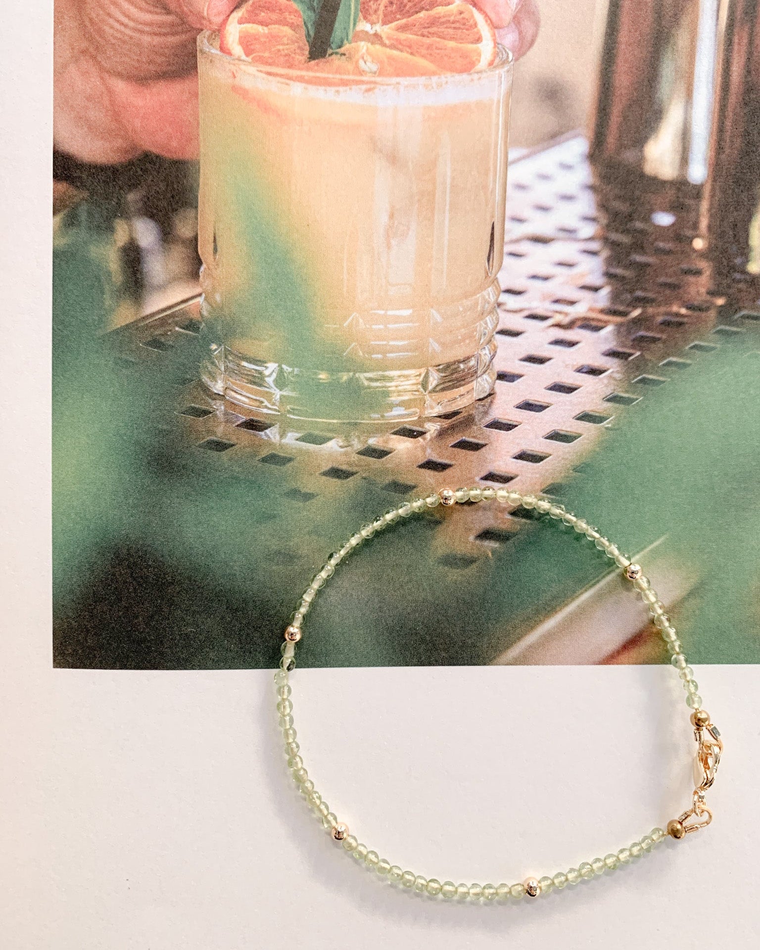 A delicate beaded bracelet with pale green beads and small gold spacer beads and clasp, displayed on a white surface below a lifestyle photo of a hand holding a cocktail with a green garnish.