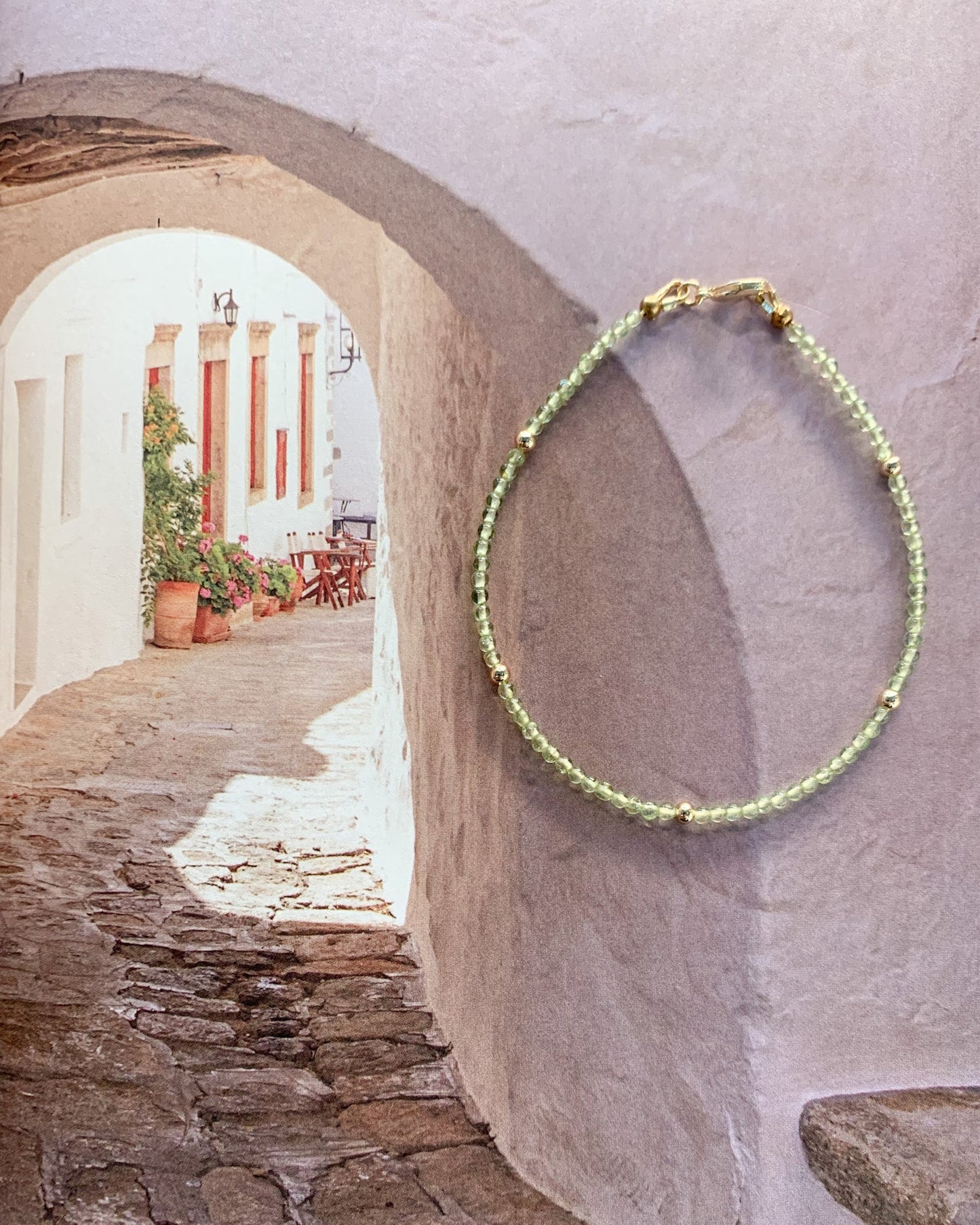 A beaded bracelet with light green peridot beads and small gold accents hangs on a textured white wall in a Mediterranean-style stone alleyway.