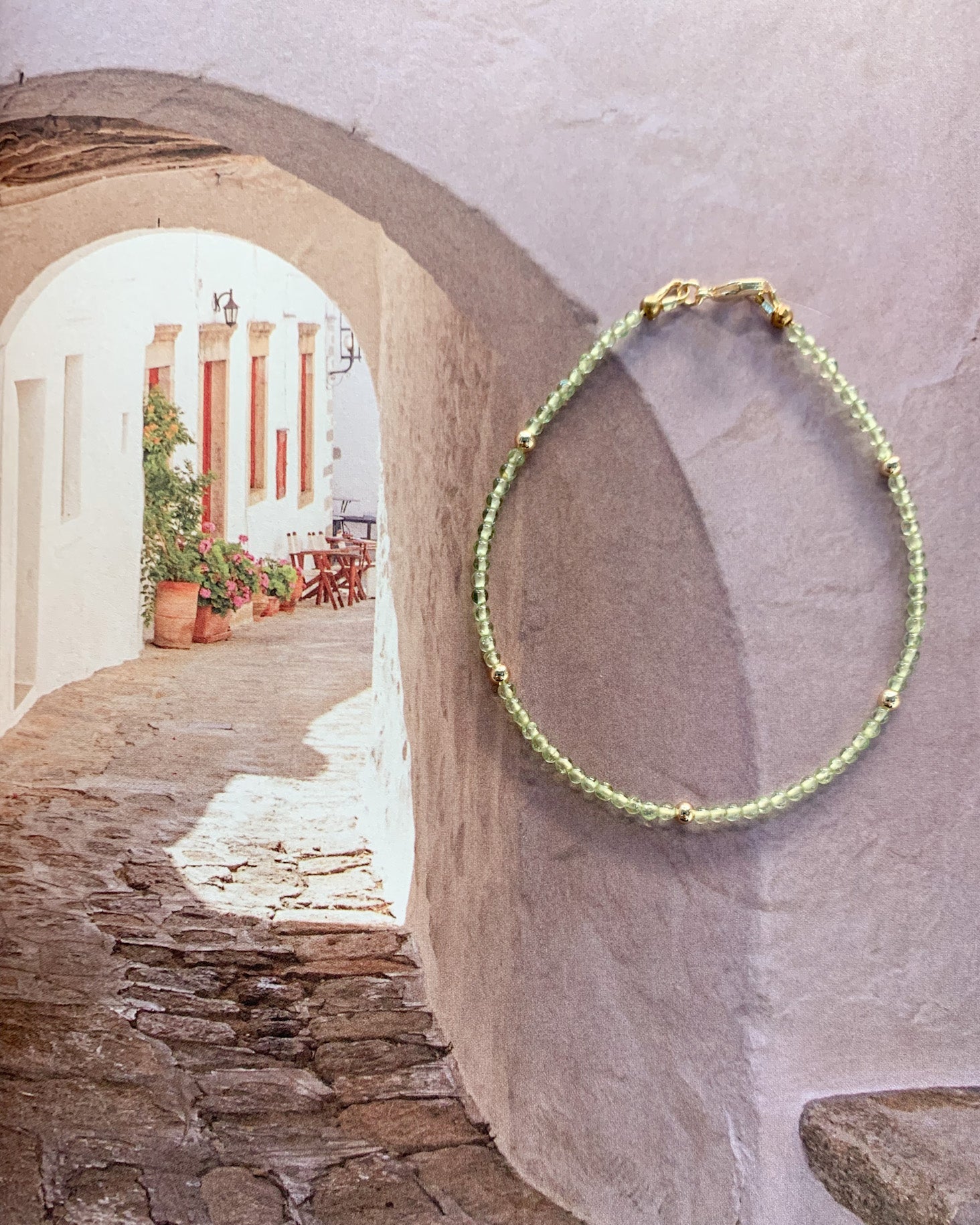 A beaded bracelet with light green peridot beads and small gold accents hangs on a textured white wall in a Mediterranean-style stone alleyway.