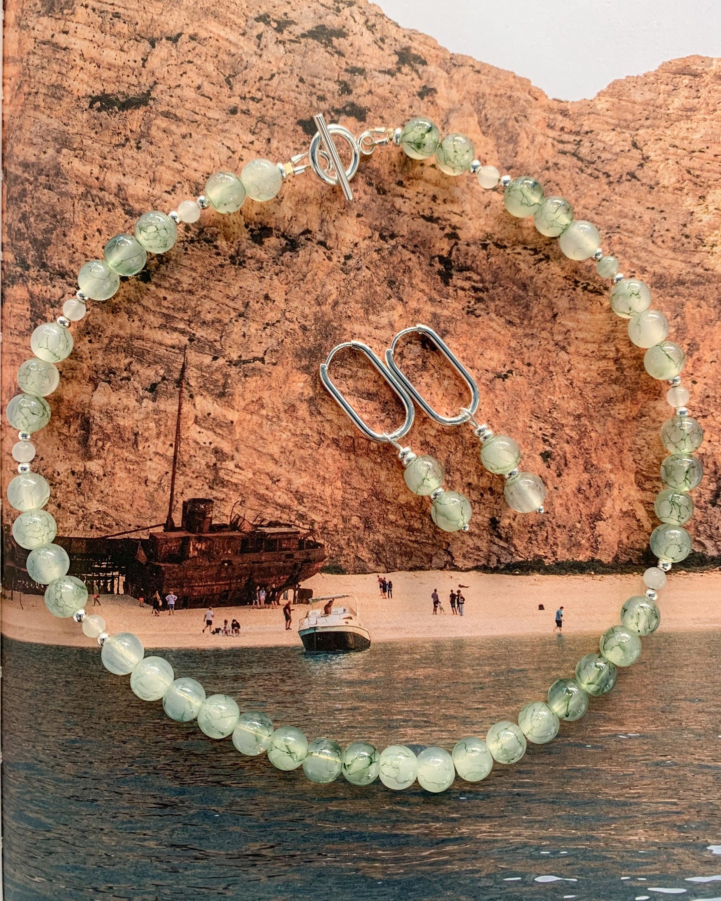 A beaded necklace and matching drop earrings with pale green translucent round beads and silver-toned metal hardware, arranged over a beach scene background featuring tan cliffs, water, and a shipwreck.