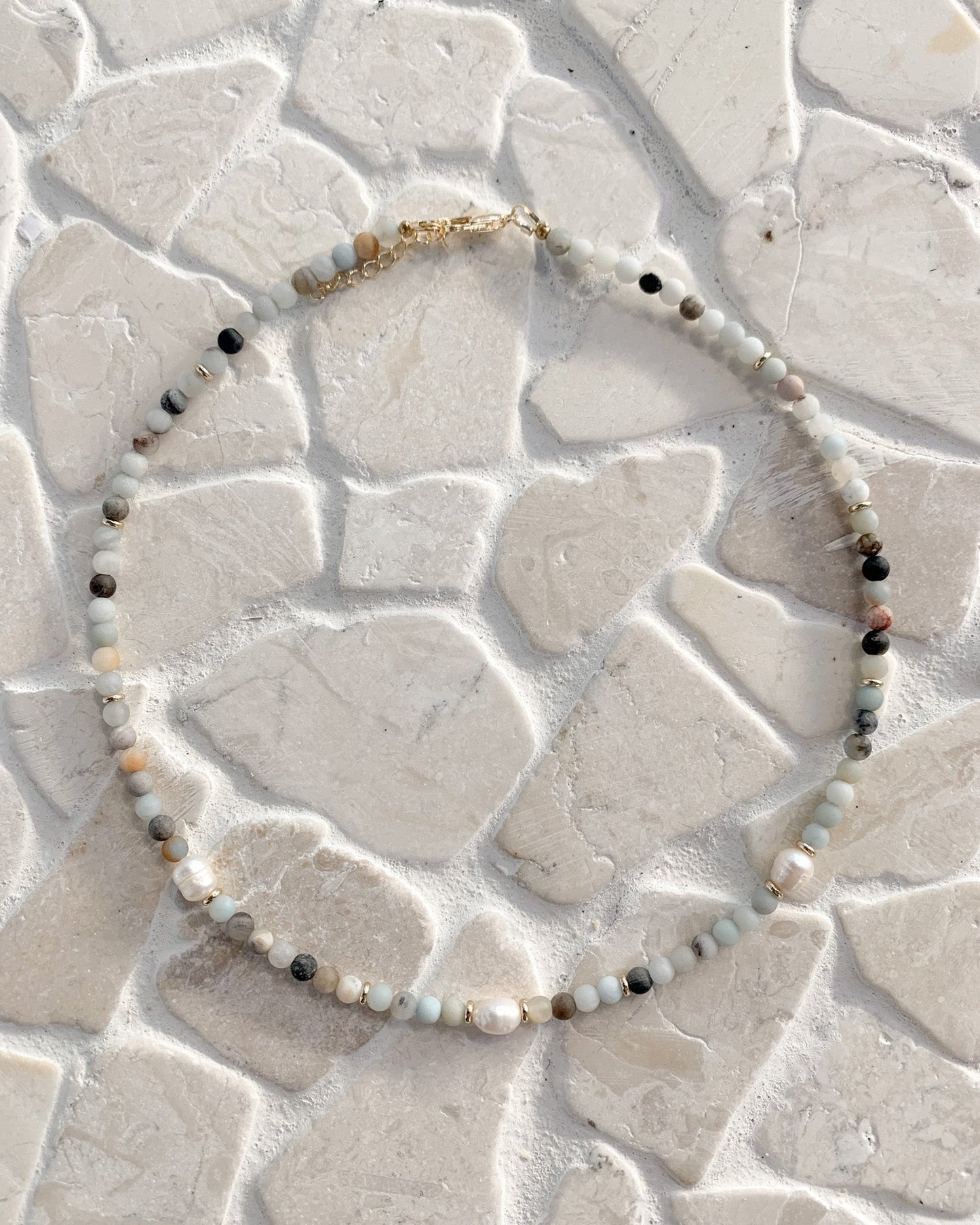 A beaded necklace with small white, gray, and black beads and a gold clasp, arranged in a circle on a textured light beige stone surface.
