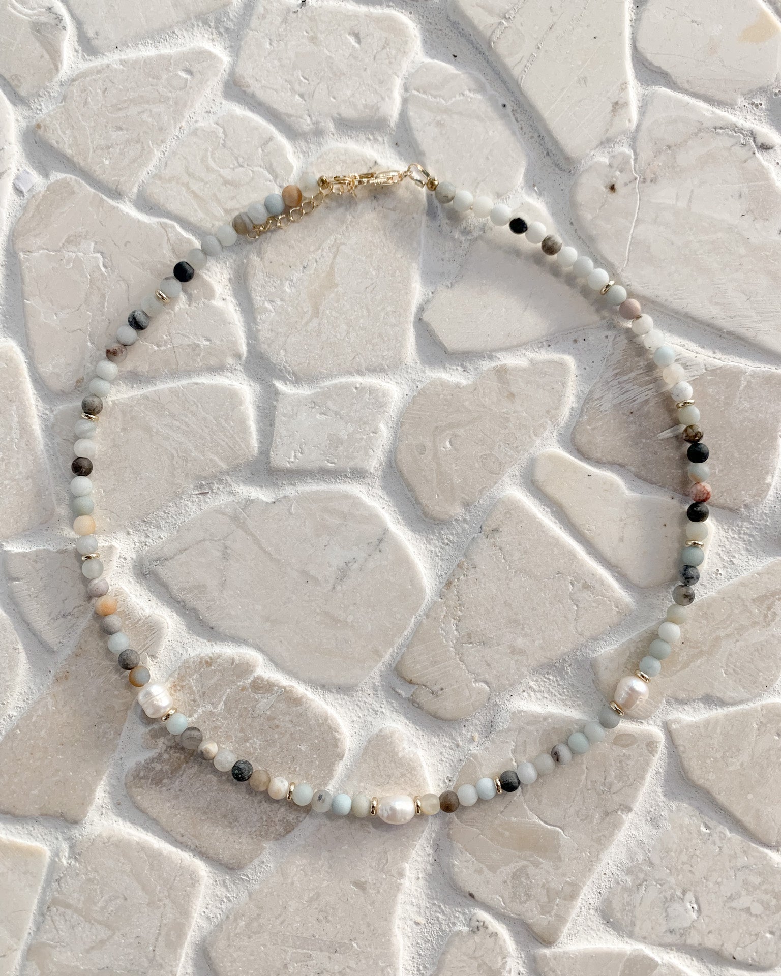 A beaded necklace with small white, gray, and black beads and a gold clasp, arranged in a circle on a textured light beige stone surface.