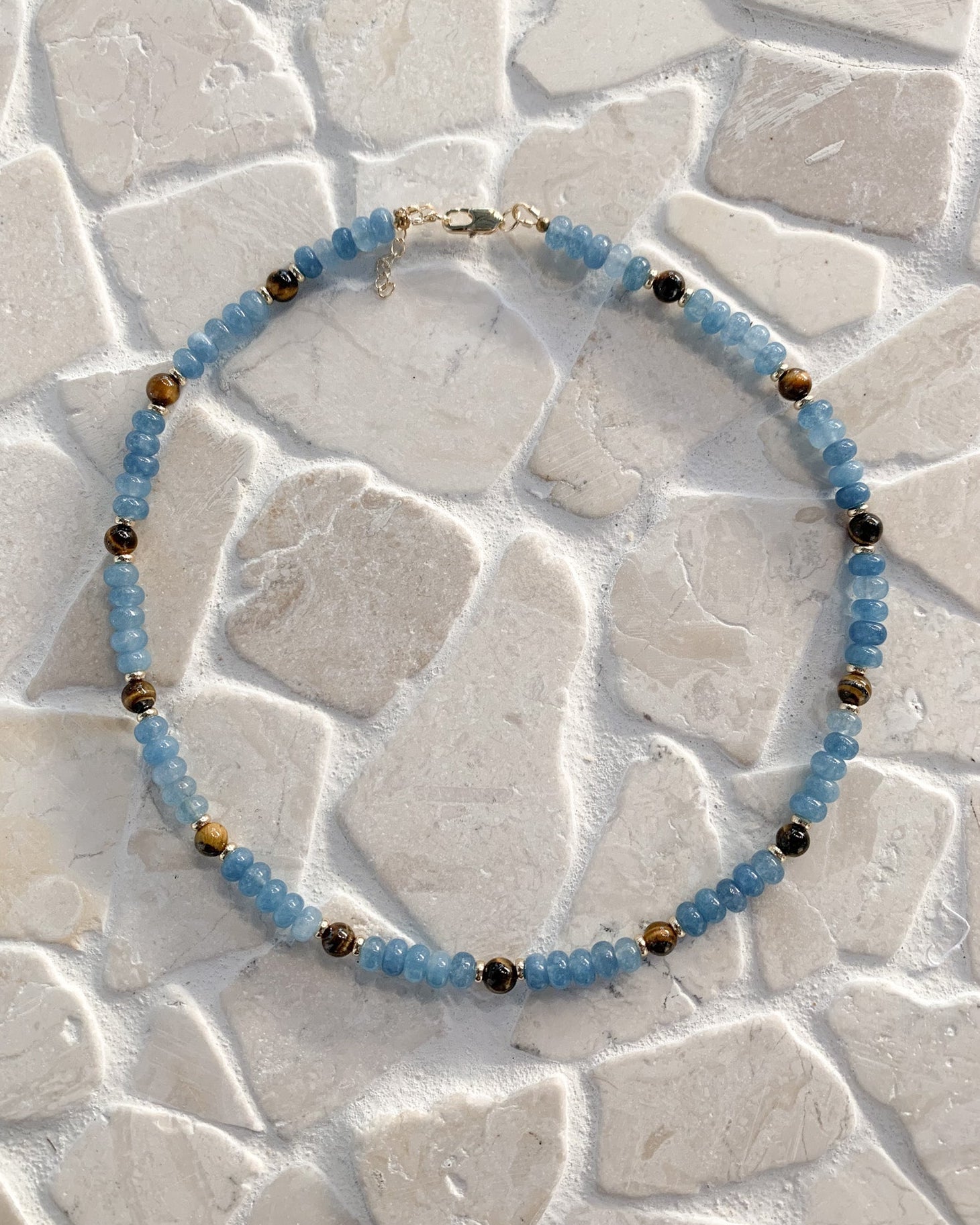 A beaded necklace with light blue faceted beads and small metallic gold spacer beads, arranged in a circle on a light beige stone tile background.