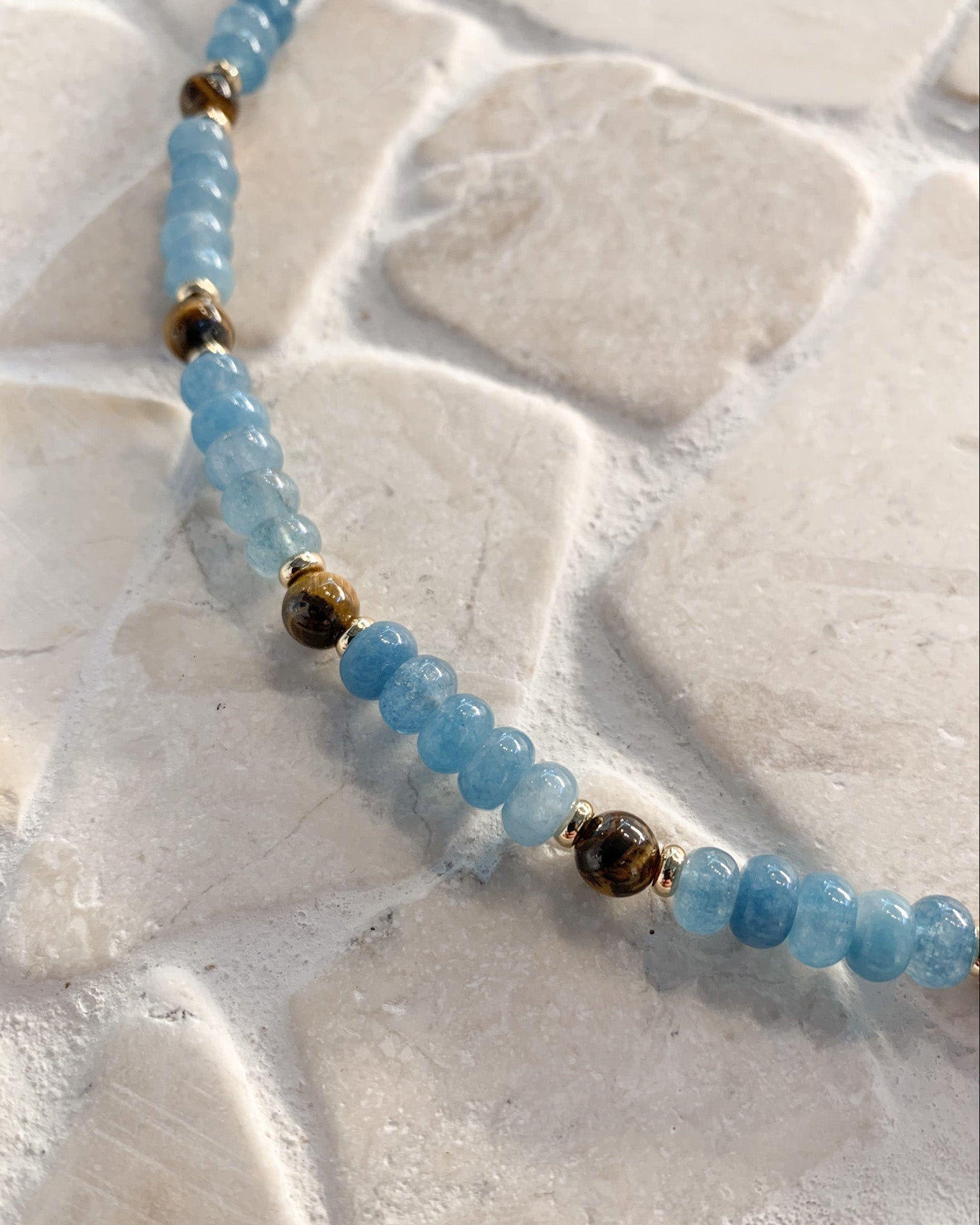 A beaded strand of light blue round beads separated by small gold spacer beads, with one larger dark brown tiger's eye accent bead, laid on a light beige stone surface.