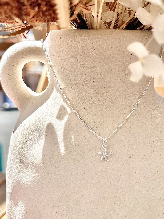 Silver starfish necklace with tiny cubic zirconia displayed on neutral ceramic vase with dried florals.