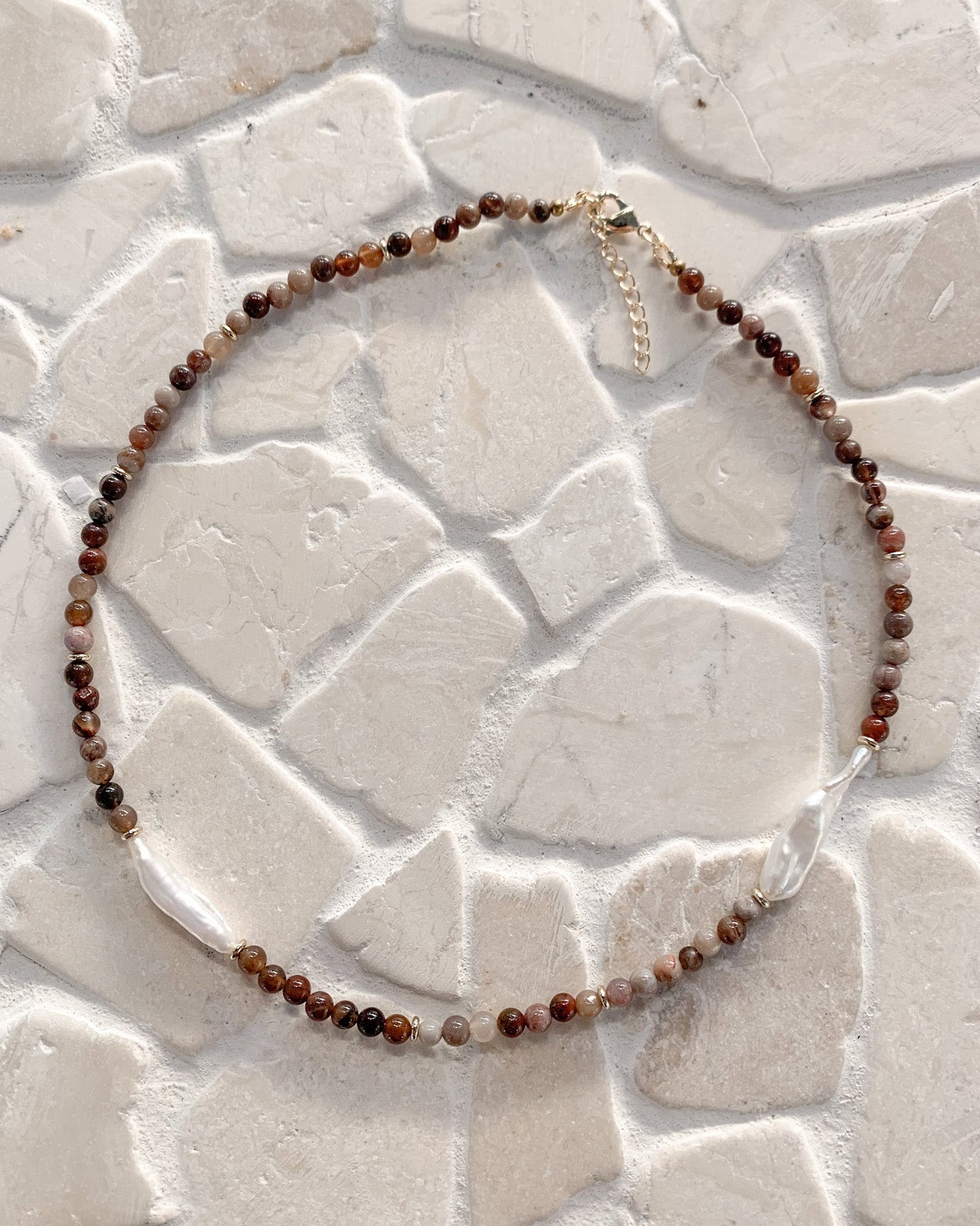 A beaded necklace made of small brown, reddish-brown, and translucent grey bamboo agate beads and freshwater pearls  with a gold-tone clasp, displayed on a light beige stone tile background.
