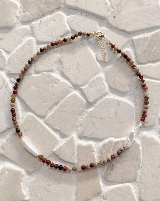 A beaded necklace made of small brown, reddish-brown, and translucent grey bamboo agate beads and freshwater pearls  with a gold-tone clasp, displayed on a light beige stone tile background.