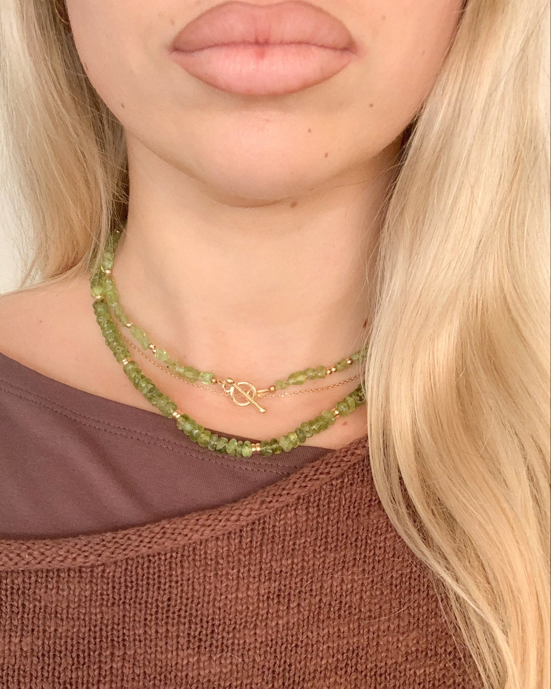 Close-up of a model wearing two layered green beaded peridot necklaces with a small gold front toggle clasp, styled over a brown knit sweater.