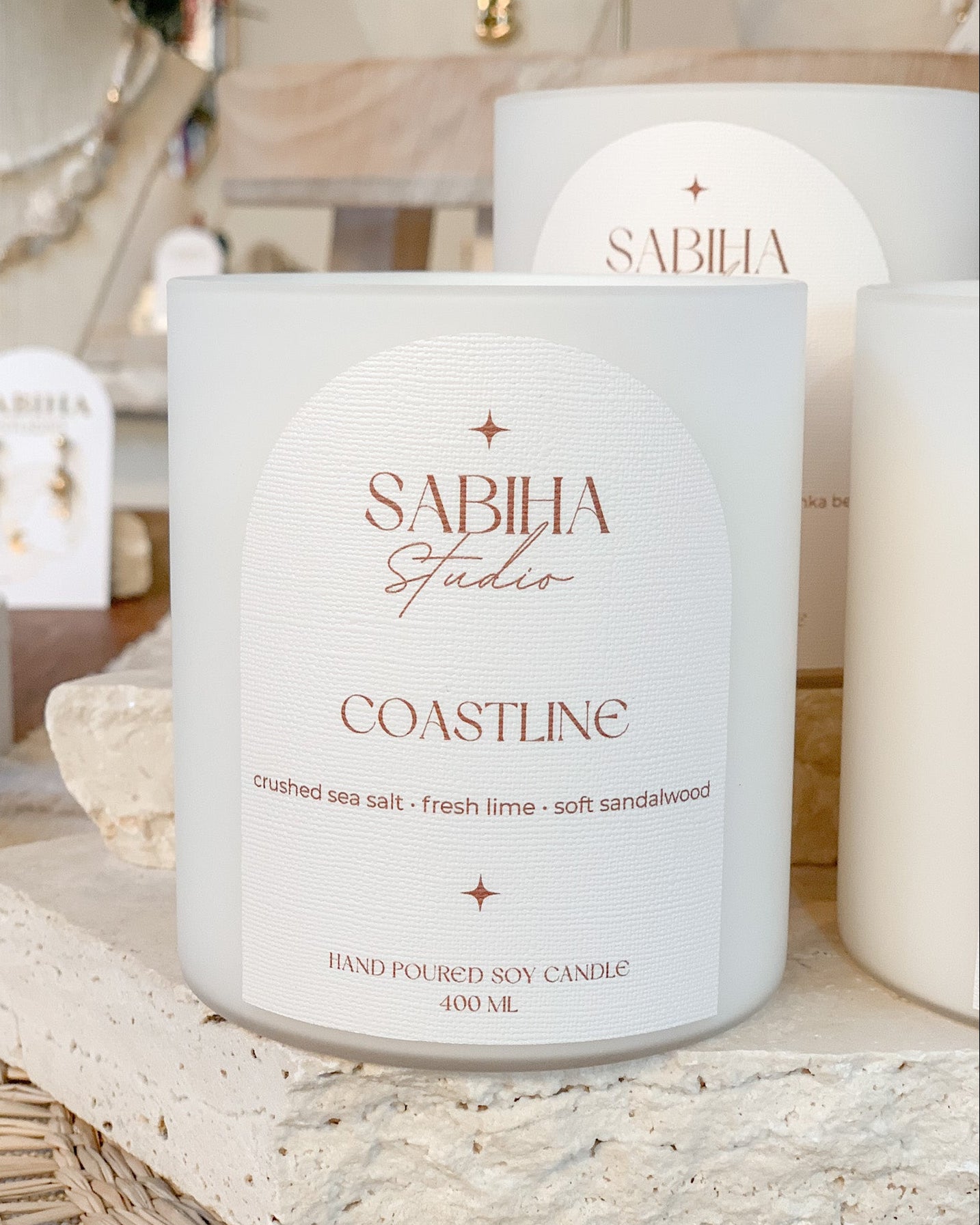 A white soy candle jar labeled 'SABIHA Studio COASTLINE' sits on a stone slab in a styled indoor setting with other candles and jewelry displays in the background.
