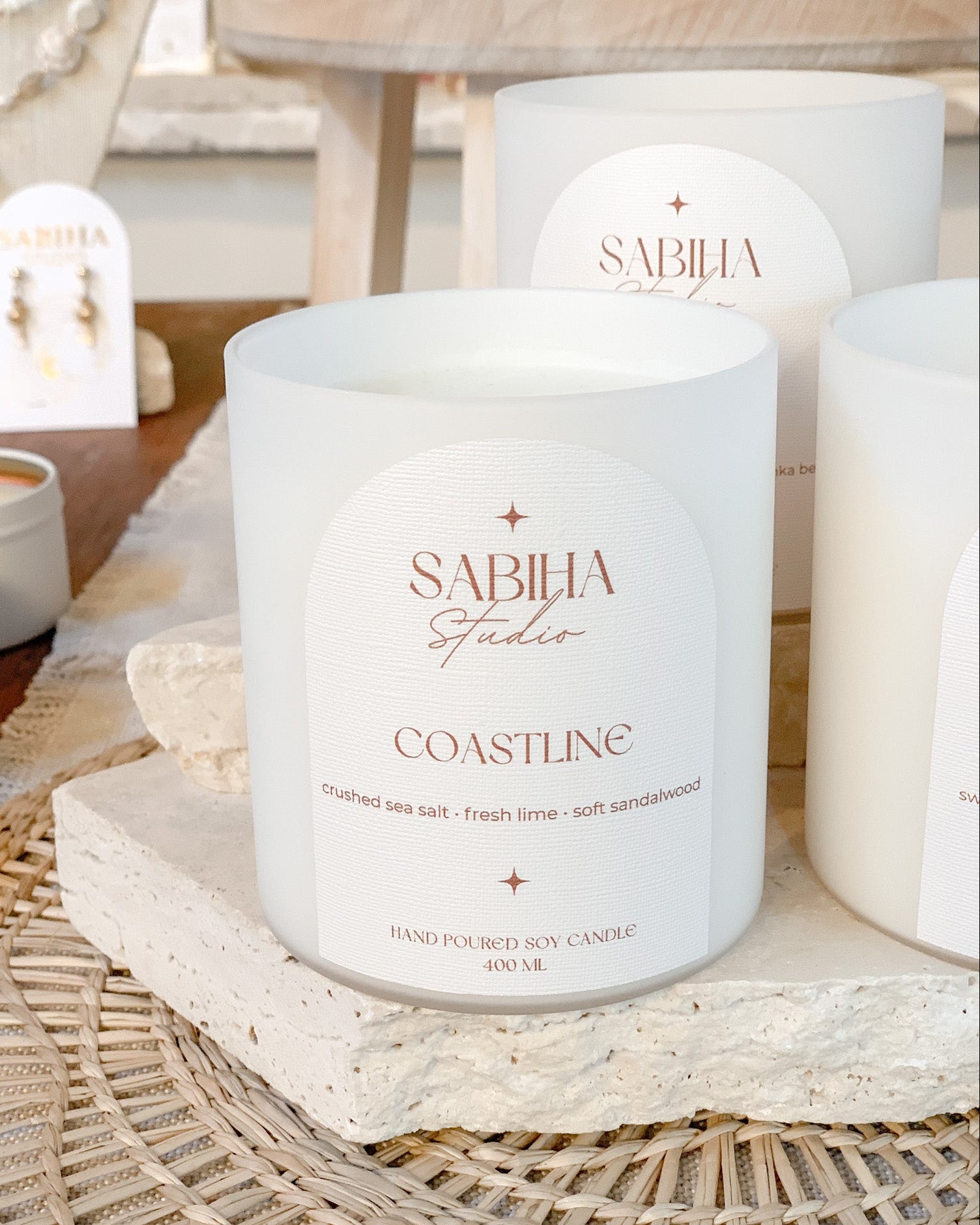 A white soy candle jar labeled 'SABHIA Studios COASTLINE' sits on a woven tray with other matching candles and a small jewelry display in a softly styled indoor setting.