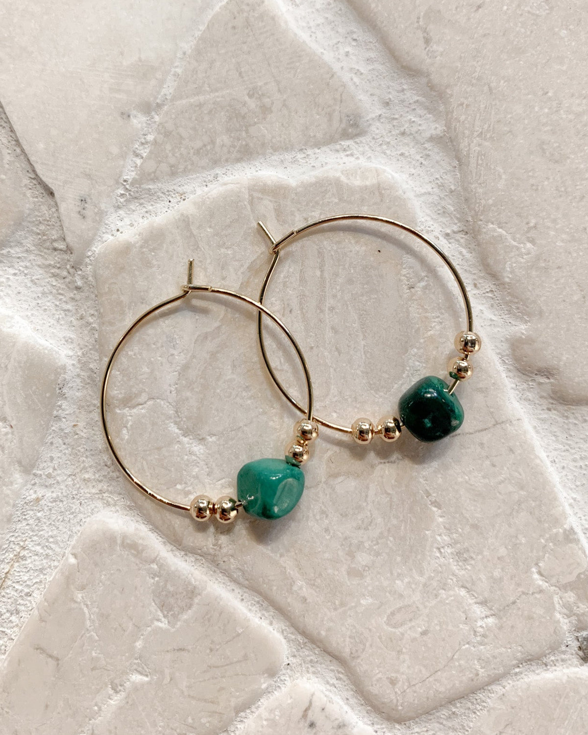Gold hoop earrings with green malachite beads, displayed on a light beige stone surface.