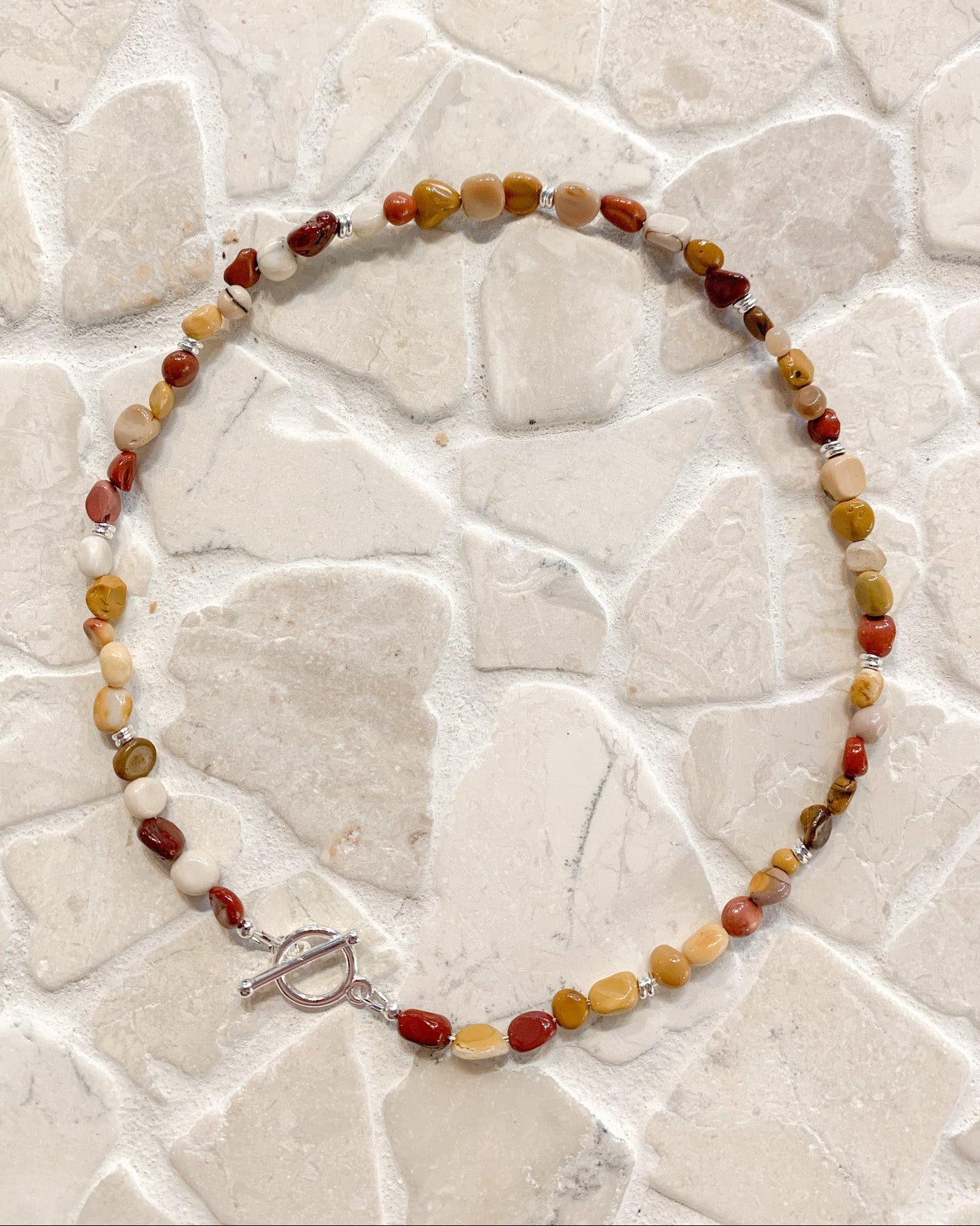 A beaded necklace made of small polished mookalite stones in red, orange, yellow, cream, and brown tones, with a silver toggle clasp, displayed on a light stone-tile surface.