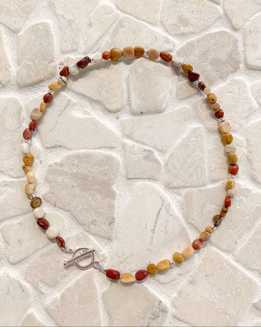 A beaded necklace made of small polished mookalite stones in red, orange, yellow, cream, and brown tones, with a silver toggle clasp, displayed on a light stone-tile surface.