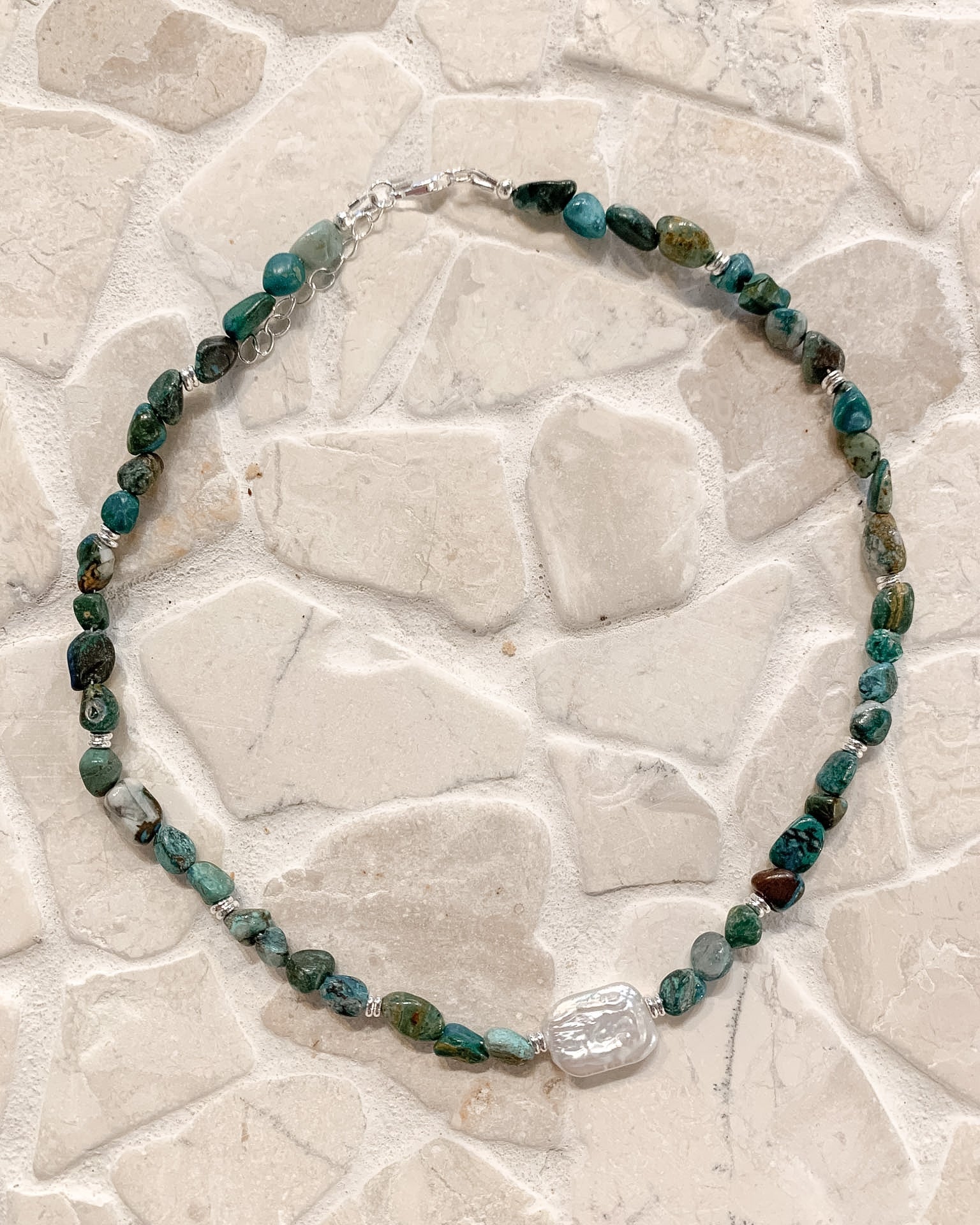 A beaded necklace made of polished green and blue stones with a clear faceted crystal pendant, displayed on a light beige stone tile background.