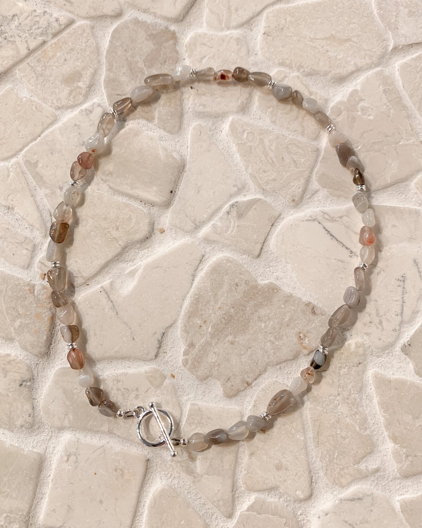 A beaded necklace made of irregular translucent gray and brown gemstone beads with a silver toggle clasp, laid flat on a light beige stone tile surface.