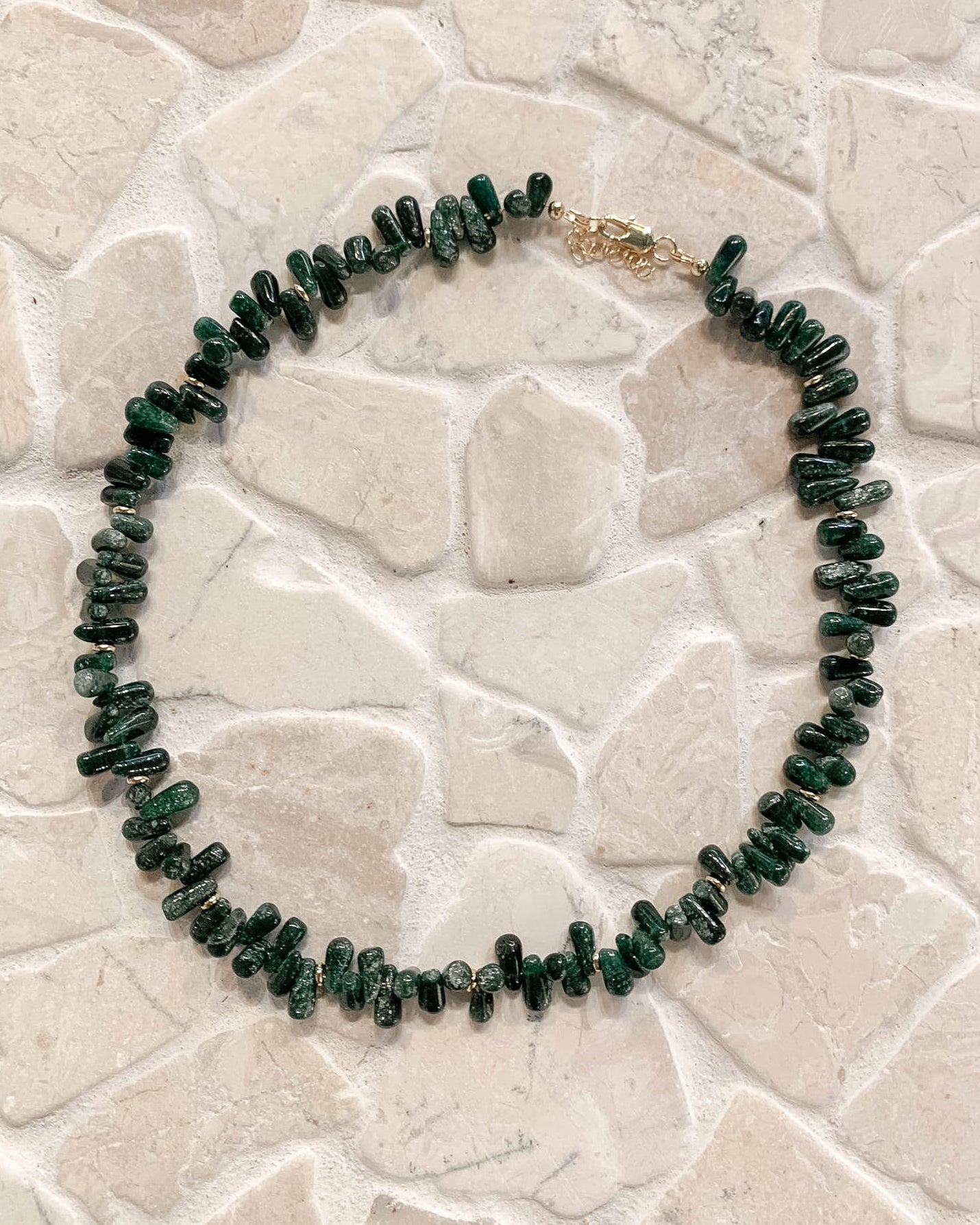 A beaded necklace made of small dark green stone chips with a gold clasp, displayed on a light beige stone tile surface.