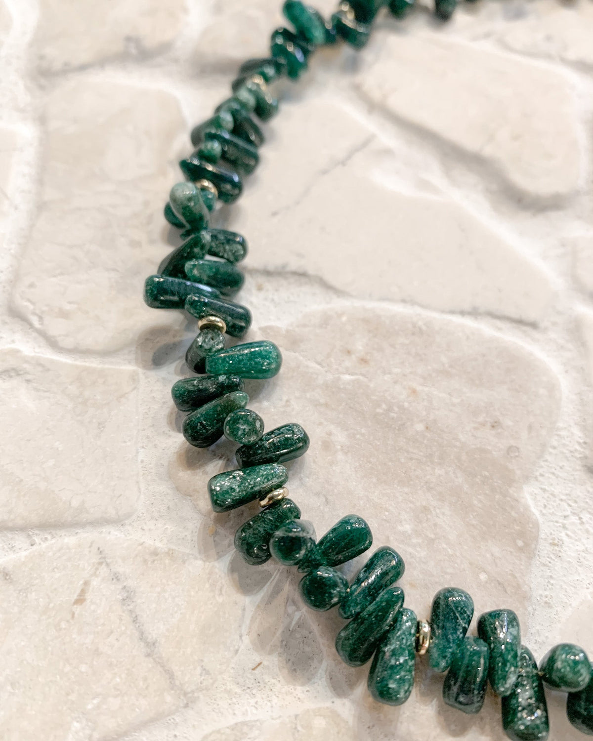A close-up of a chunky beaded necklace made of polished green stone chips, laid out on a light beige stone surface.