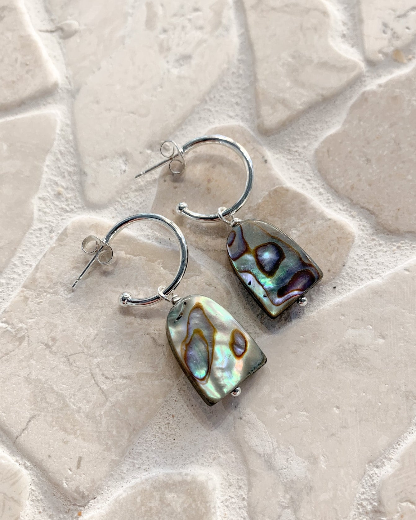 A pair of silver hoop earrings with irregular abalone shell pendants, displayed on a light beige stone tile background.