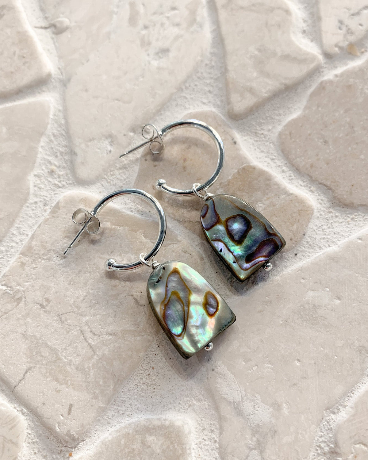 A pair of silver hoop earrings with irregular abalone shell pendants, displayed on a light beige stone tile background.
