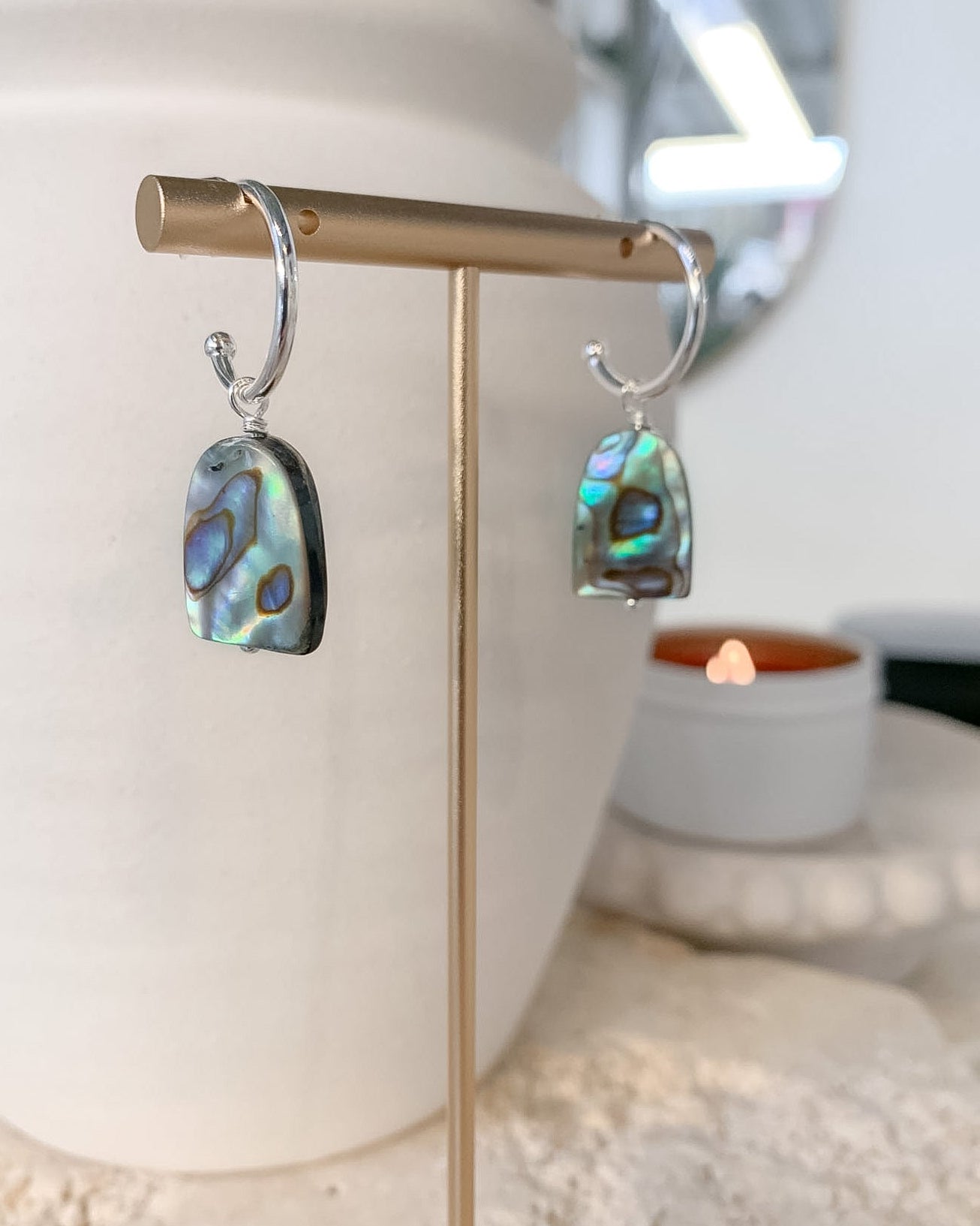A pair of silver hoop earrings with irregular iridescent abalone shell pendants, displayed on a gold stand in a styled indoor setting with a white planter and a lit candle in the background.