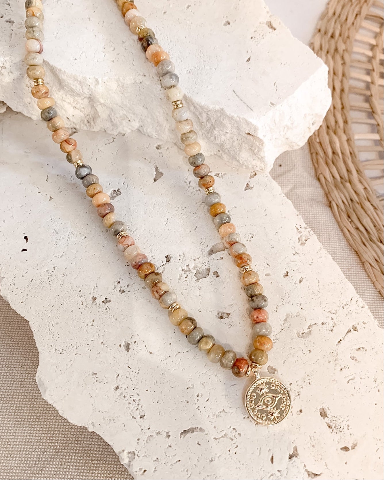 A beaded necklace with earthy marbled beads in beige, brown, orange, and gray, featuring a gold-toned coin pendant, displayed on a textured white stone slab.