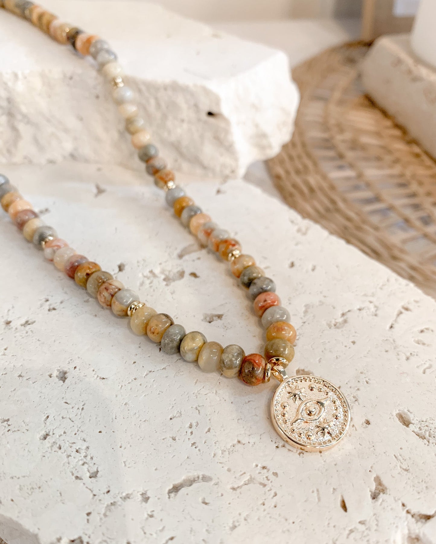A beaded necklace with marbled stone beads in earthy tones and a gold-toned circular pendant, styled on a textured white stone surface.