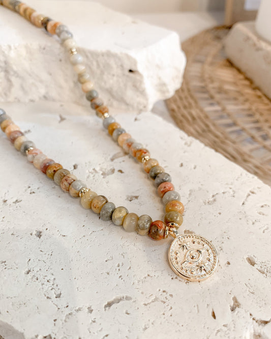 A beaded necklace with marbled stone beads in earthy tones and a gold-toned circular pendant, styled on a textured white stone surface.