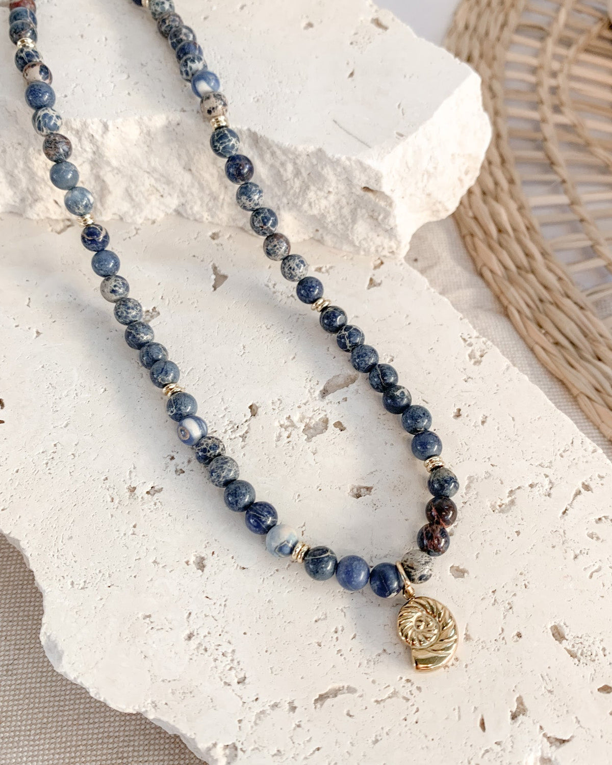 A beaded necklace with dark blue and grey marbled sodalite beads, gold spacer beads, and a small gold skull pendant, displayed on a white stone prop.