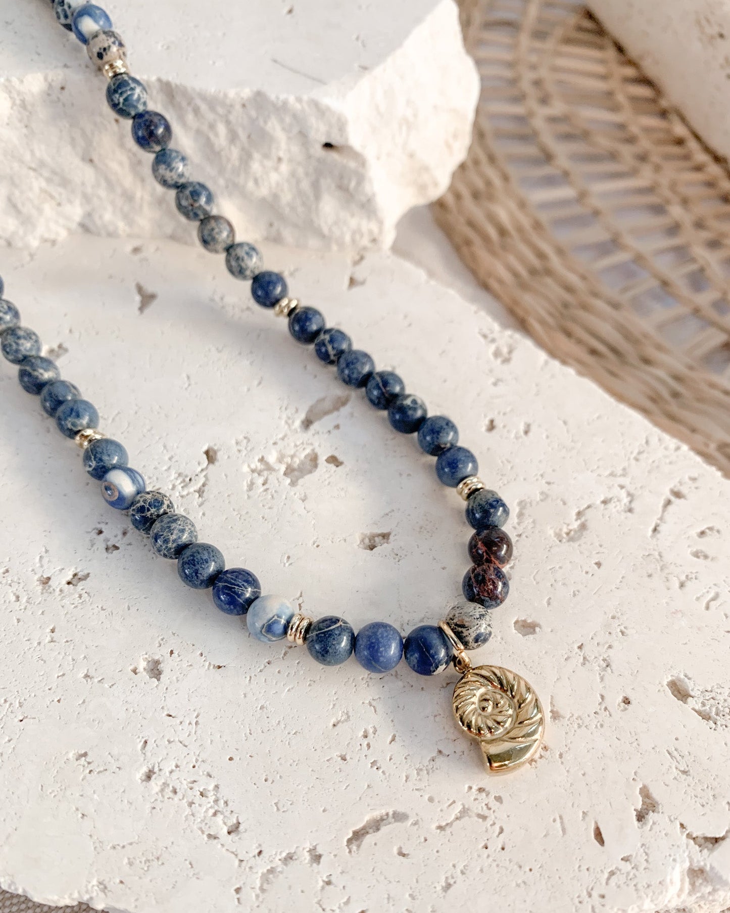 A beaded necklace with dark blue and white sodalite round beads, small gold spacer beads, and a gold-toned spiral shell pendant, displayed on a textured white stone slab.