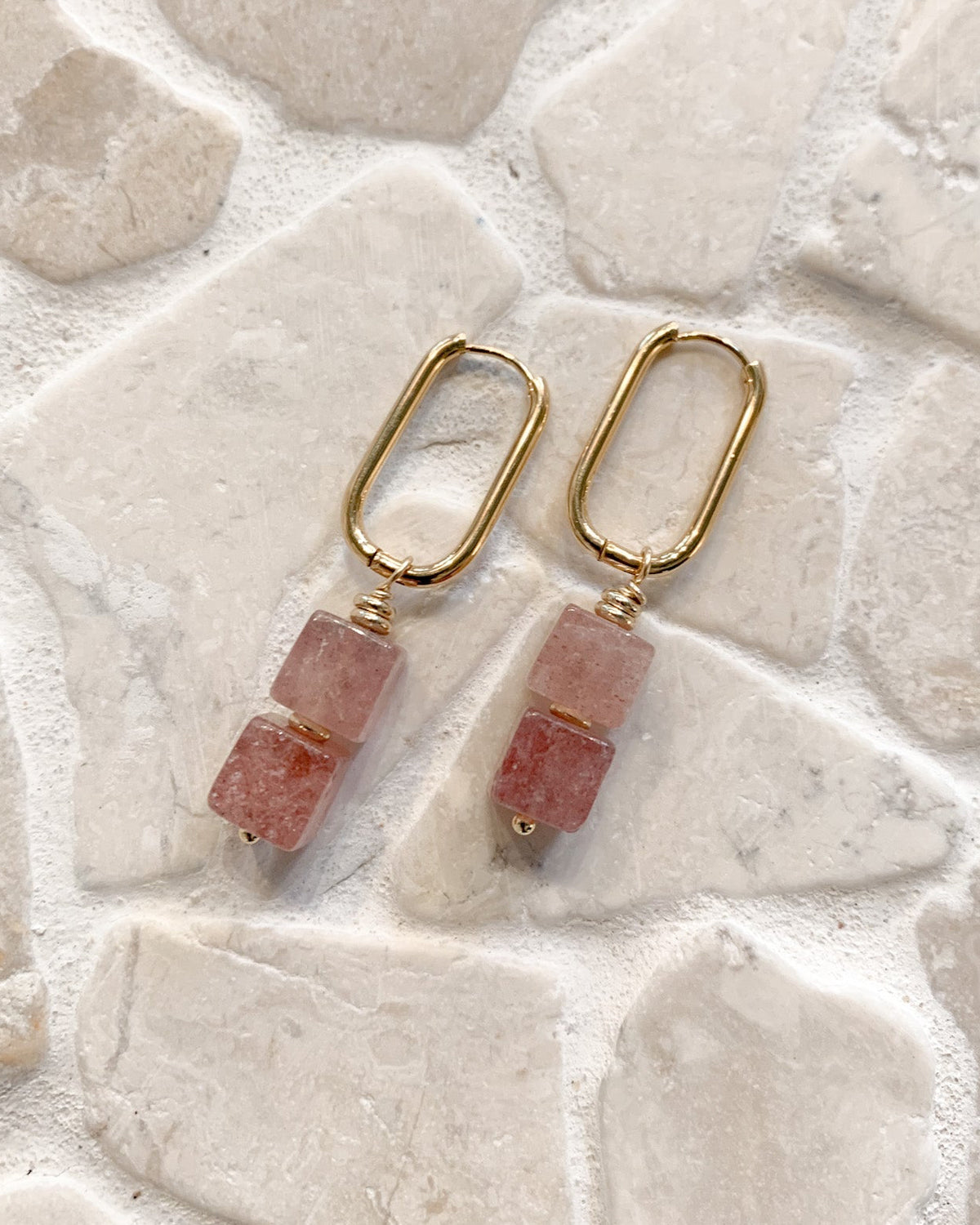 A pair of gold drop earrings with two stacked pink stone beads, displayed on a light beige stone tile background.