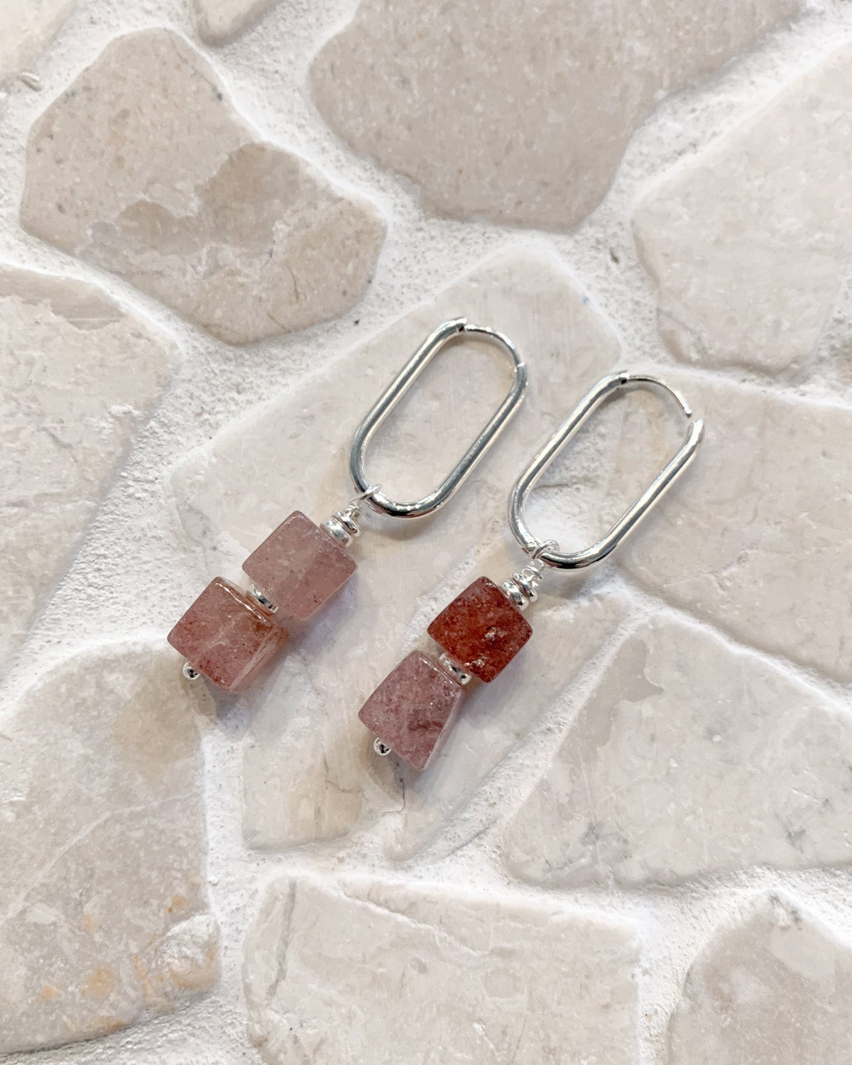 A pair of drop earrings with rectangular pinkish-red stone beads and silver-toned metal hooks, displayed on a light stone tile surface.