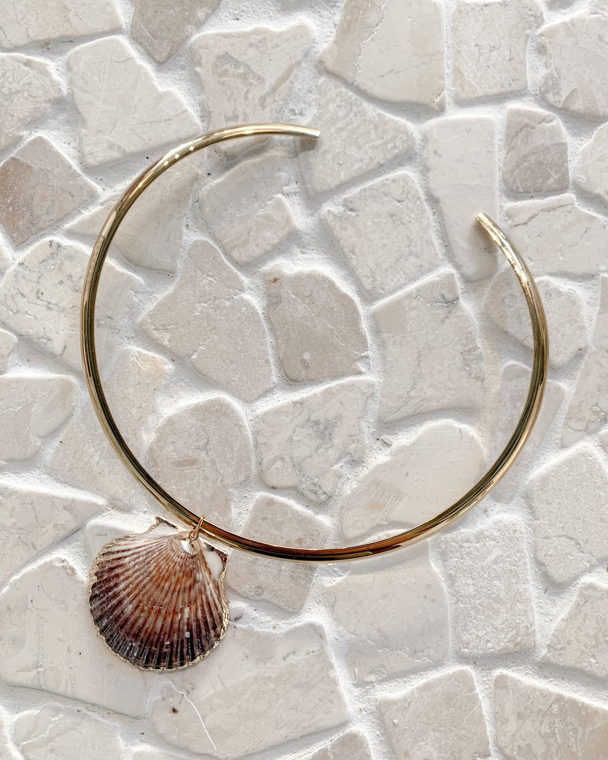 A gold-toned open hoop earring with a dangling scallop shell charm, displayed on a light beige stone mosaic tile background.
