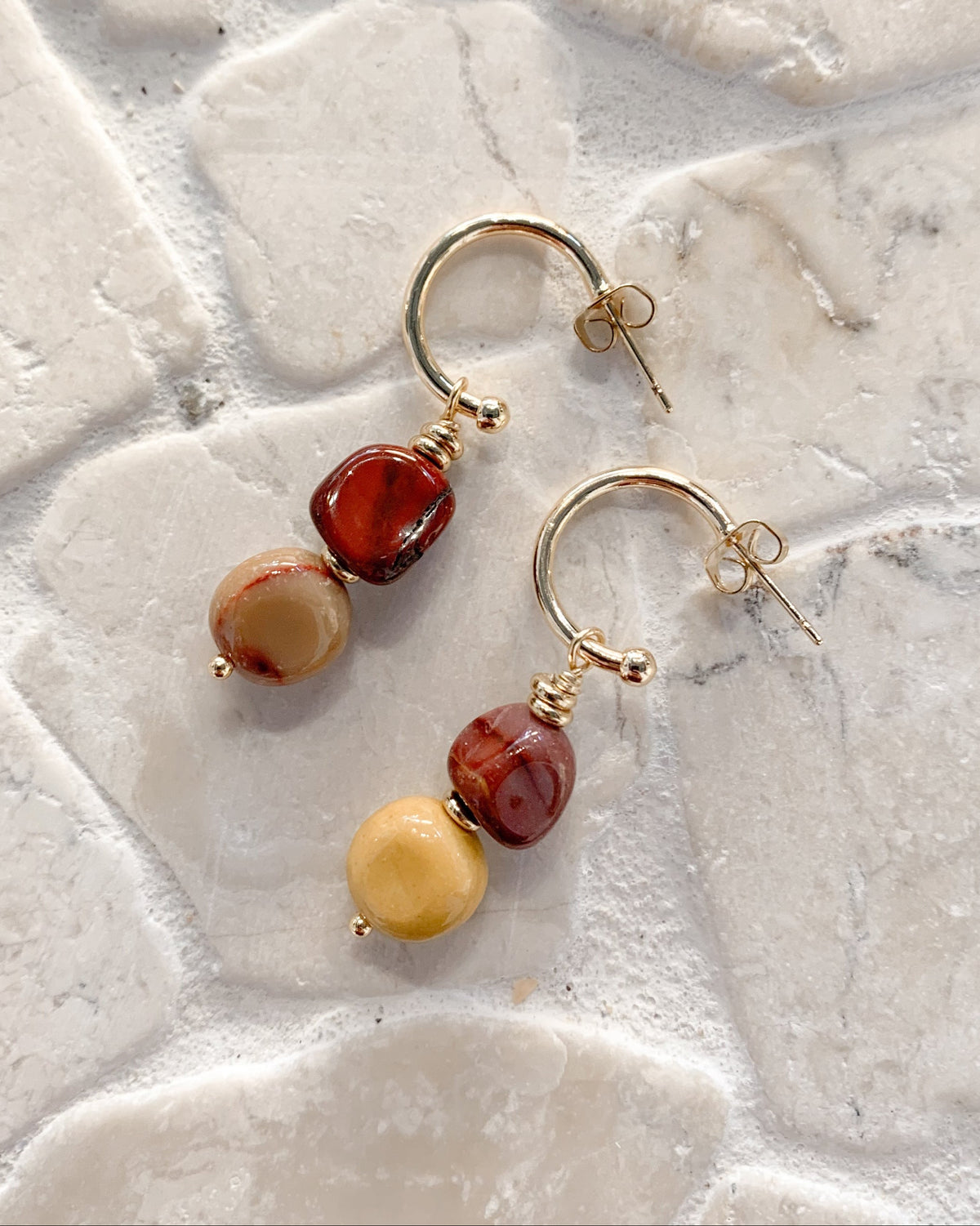 A pair of gold hoop earrings with dangling beads, displayed on a light-colored stone tile surface. The earrings feature one dark reddish-brown bead, one marbled red and brown bead, and one smooth pale yellow bead.