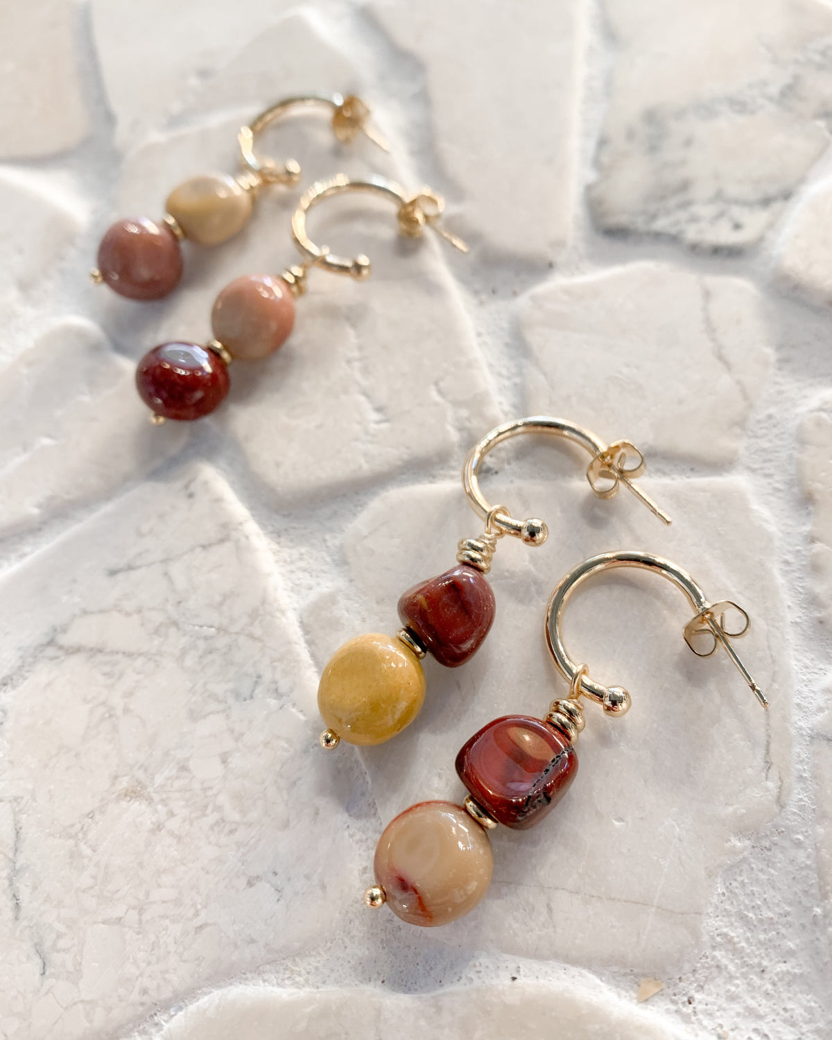 A pair of gold hoop dangle earrings with three beaded drops in earthy tones of red, yellow, and beige, displayed on a textured white stone surface.