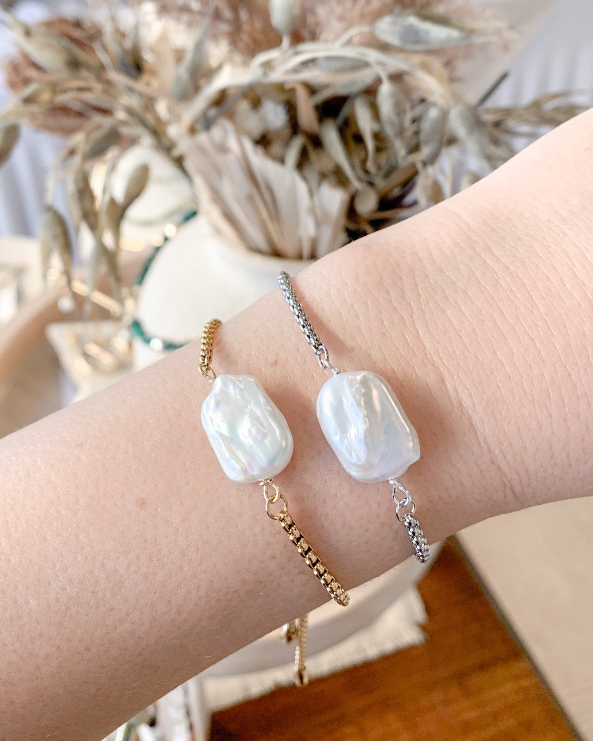 Two delicate bracelets worn on a person's wrist, featuring large white baroque pearls on gold and silver chains, set against a softly blurred boho-style background with dried pampas grass.