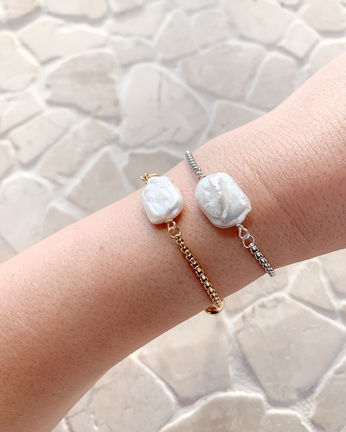 Two delicate charm bracelets worn on a person's wrist, featuring irregular white baroque pearls and gold, silver, and rose gold chains, set against a light stone tile background.