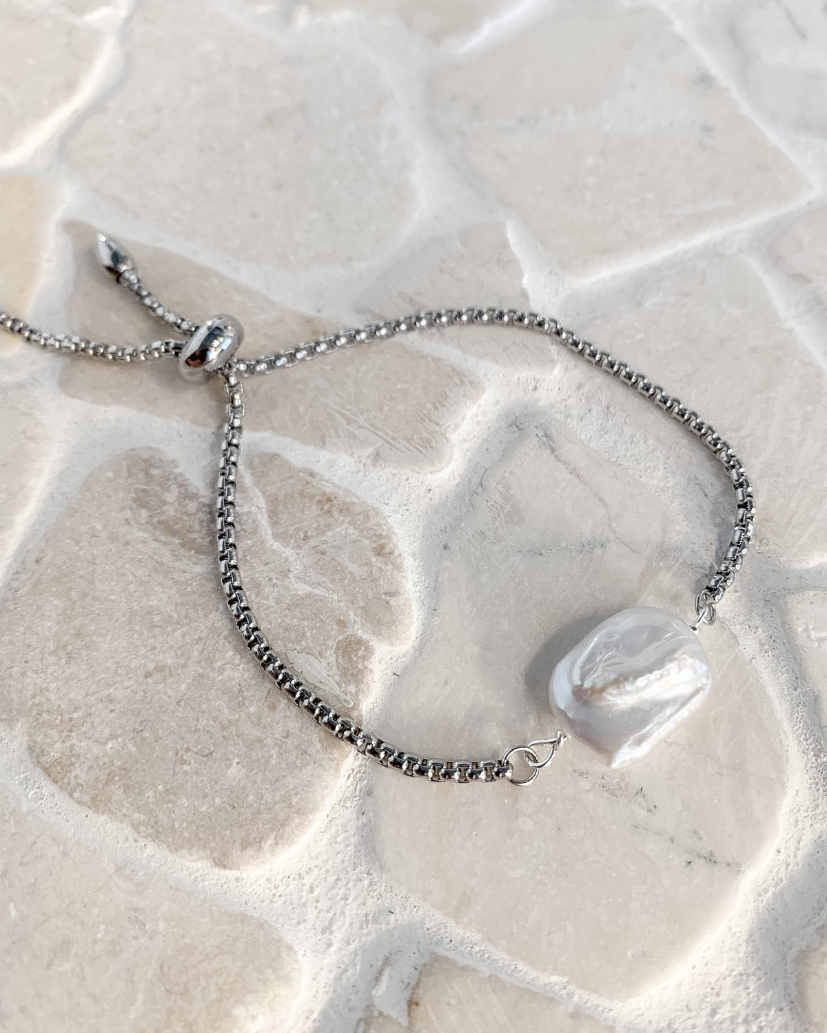 A silver chain bracelet with an adjustable sliding bead closure and a white irregular shell pendant, displayed on a light beige stone surface.
