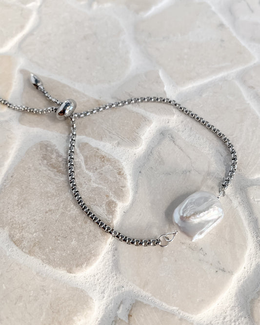 A silver chain bracelet with an adjustable sliding bead closure and a white irregular shell pendant, displayed on a light beige stone surface.