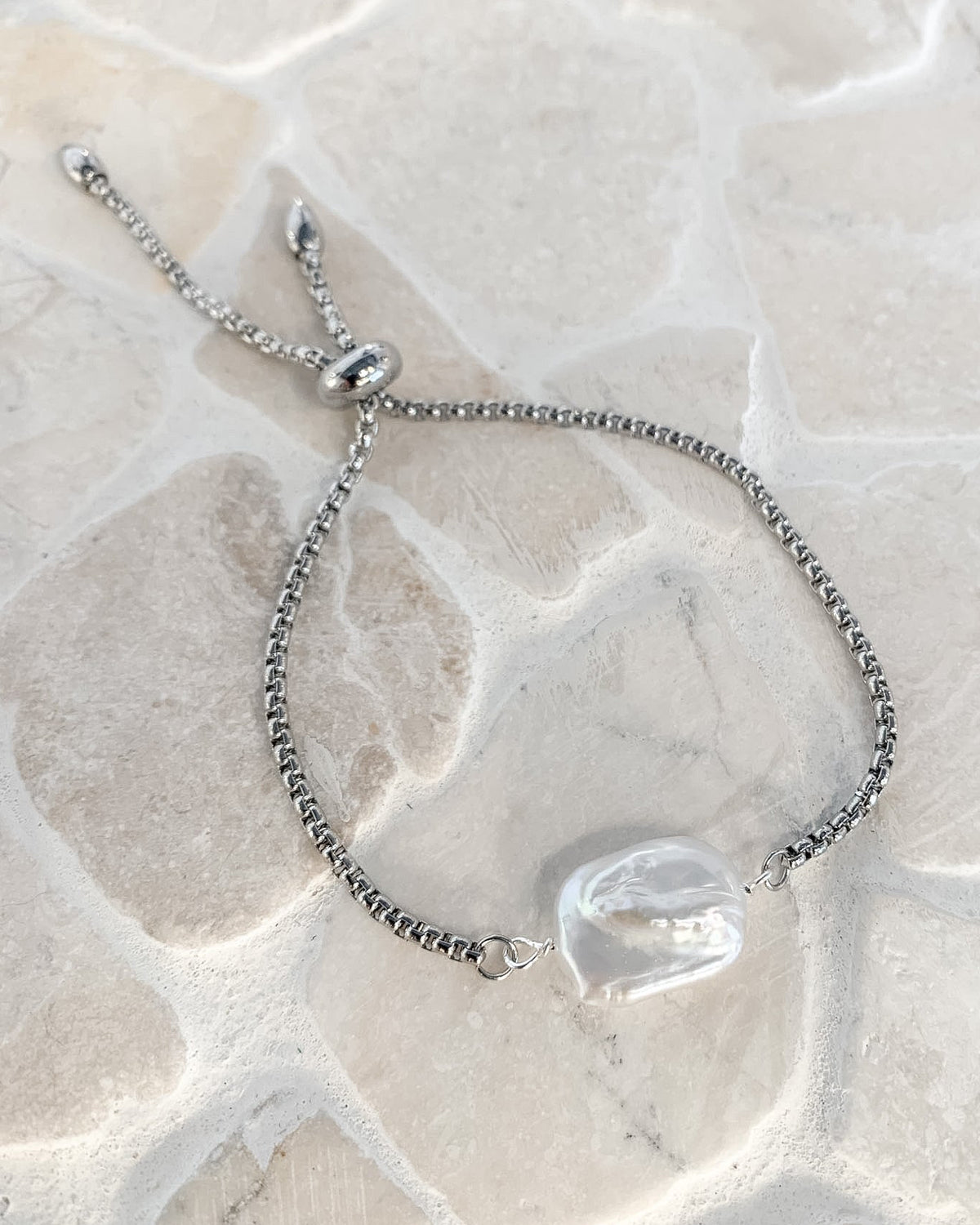 A silver adjustable rope bracelet with a large, irregular clear shell charm, displayed on a light beige stone surface.