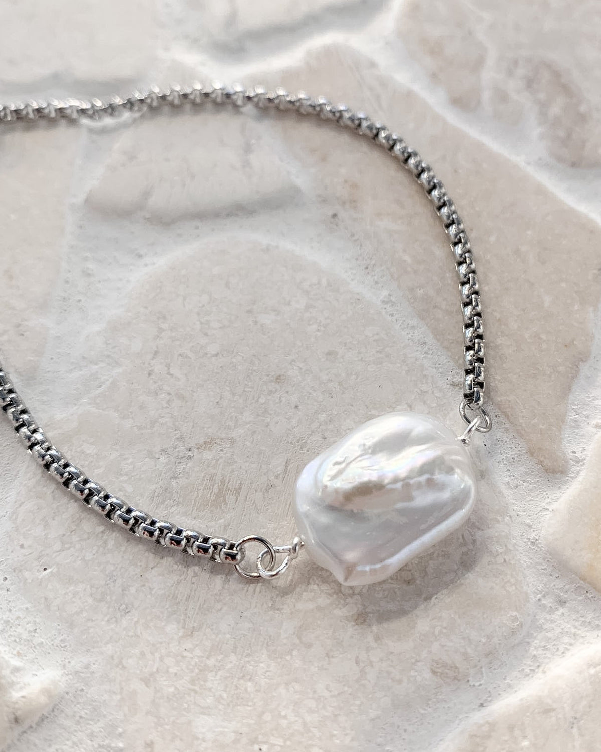 A silver chain anklet featuring a large, irregular baroque pearl pendant, displayed on a light beige stone tile surface.