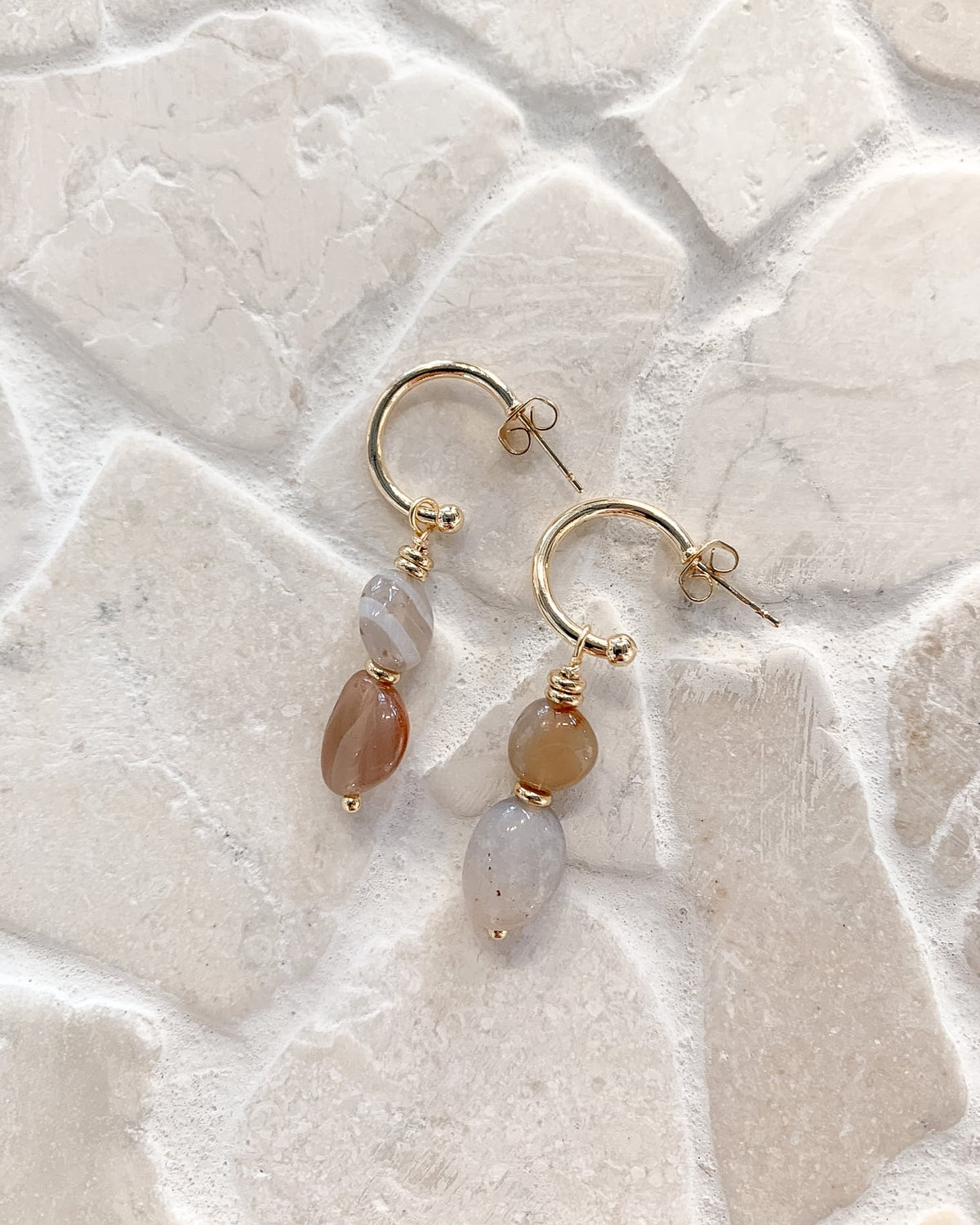 A pair of gold hoop earrings with dangling teardrop-shaped beads, one in a warm peachy-brown stone and the other in a milky translucent pale stone, displayed on a light beige stone tile background.