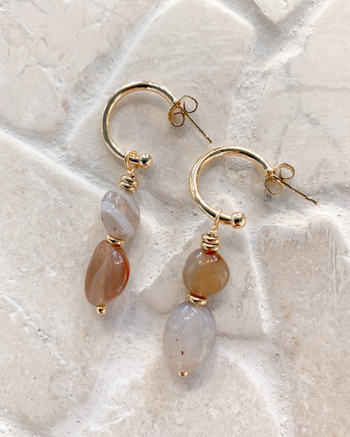 A pair of gold hoop earrings with dangling teardrop beads, one in translucent white-gray and one in warm amber-brown, displayed on a light beige stone surface.