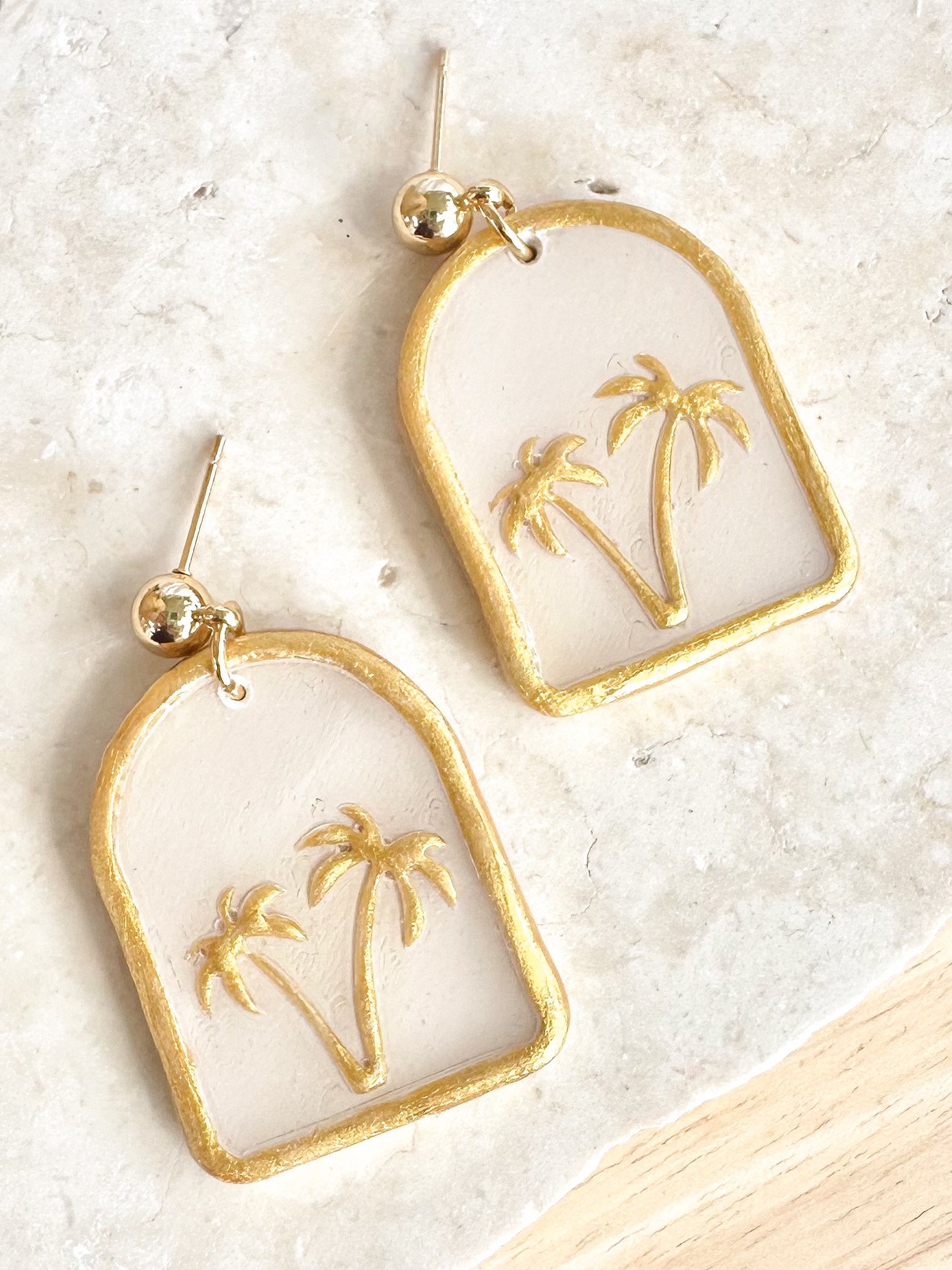 Gold and white earrings with palm tree design on a light background