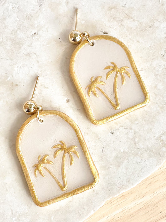 Gold and white earrings with palm tree design on a light background