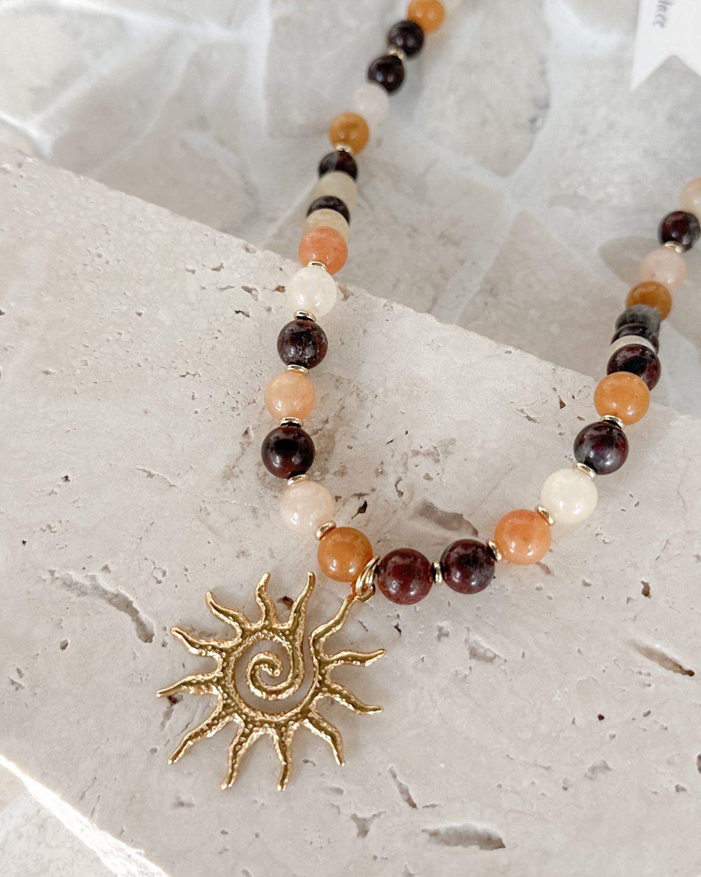 A beaded necklace with a gold sun pendant, displayed on a light stone slab over a mosaic tile background. The beads are a mix of amber, dark brown, cream, and black.