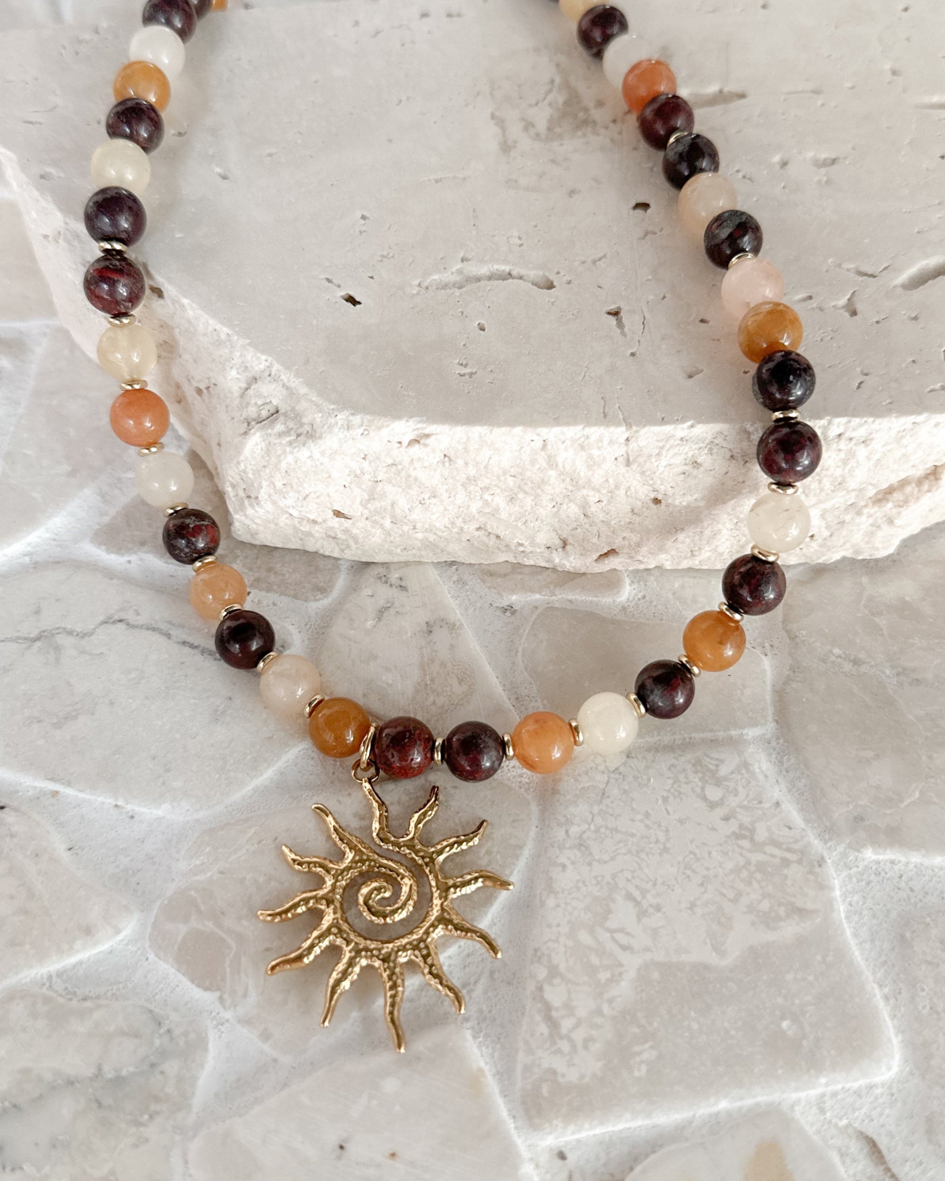 A beaded necklace with dark brown, amber, and cream beads, featuring a gold sun pendant, displayed on a textured stone slab.