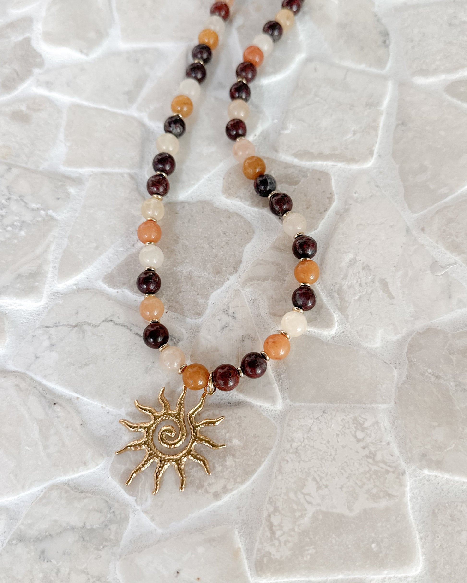 A beaded necklace with dark brown, orange, and cream beads, featuring a gold sun pendant, displayed on a light stone tile surface.