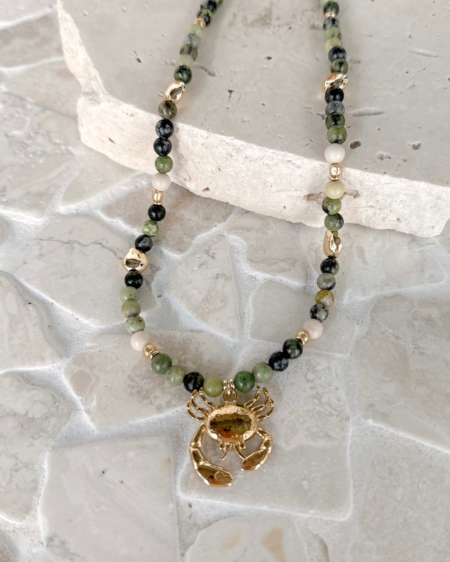 A beaded necklace with green, black, and white beads, featuring a gold-toned lobster clasp and a gold-toned lobster claw pendant, displayed on a light gray stone surface.