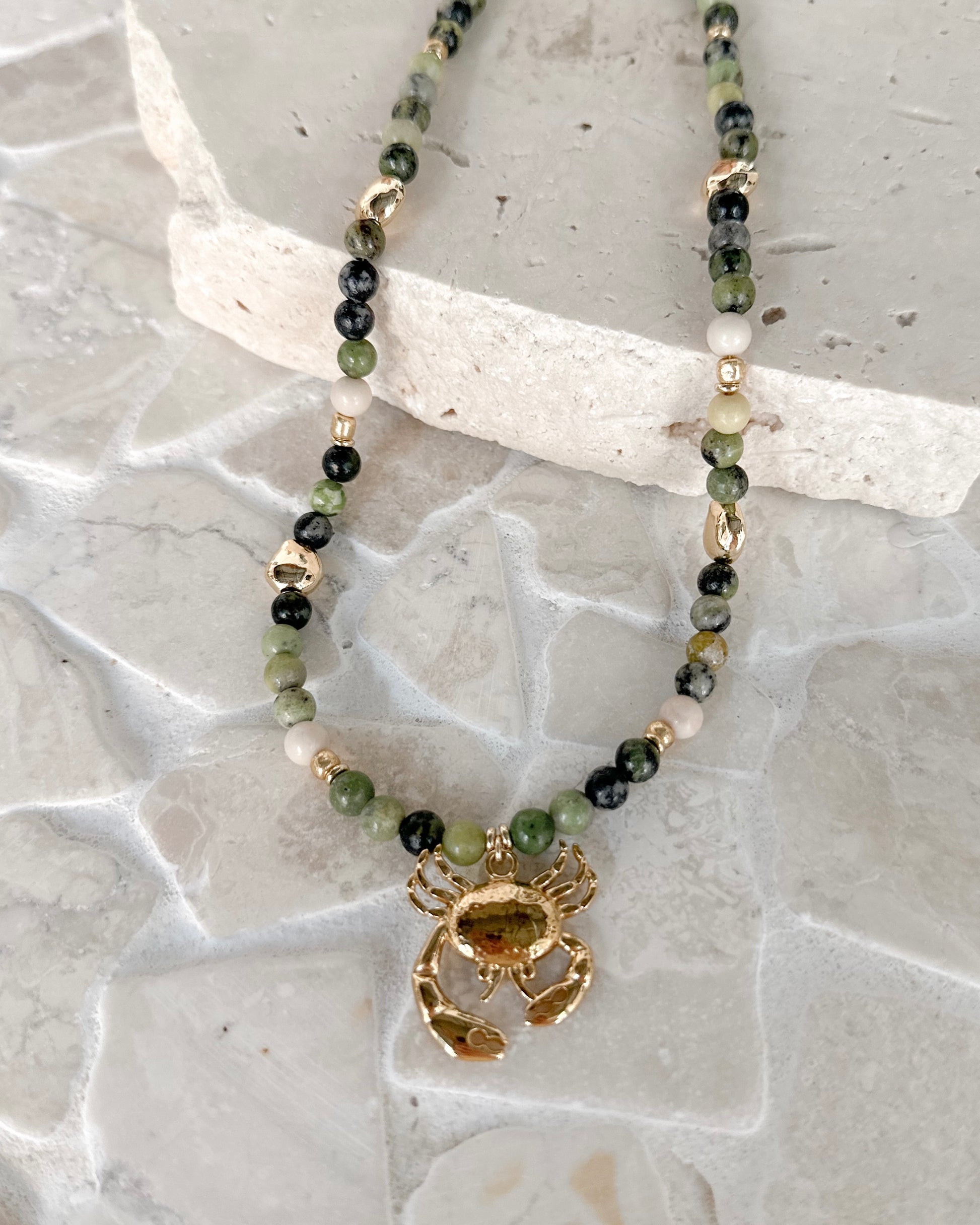 A beaded necklace with green, black, and white beads, featuring a gold-toned lobster clasp and a gold-toned lobster claw pendant, displayed on a light gray stone surface.